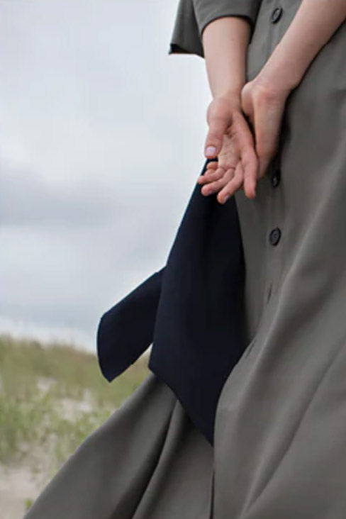 The back of a figure standing near the shore of a beach, hands clasped behind her back, wearing a long flowing grey button back dress