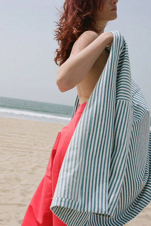woman in profile on the beach in low breaking waves - wearing bright orange/red balloon pants and a blue and white striped wrap in her arms