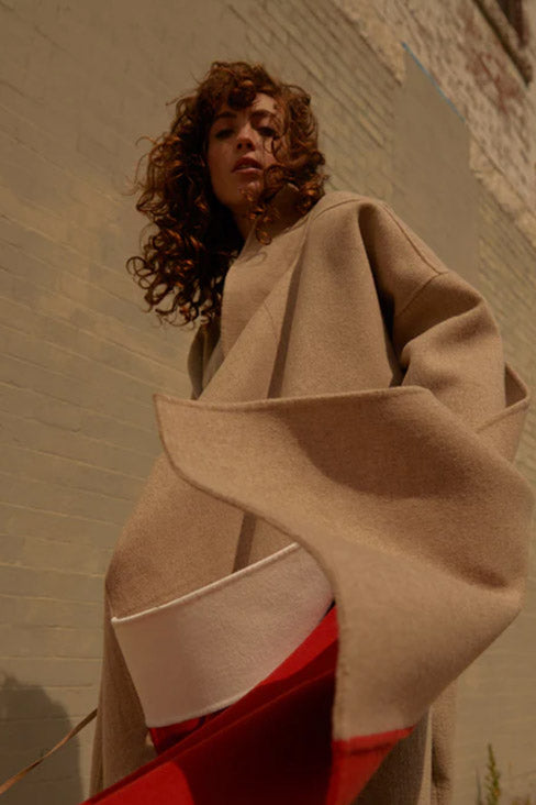 woman with long hair in front of a multi-textured beige and tan brick wall wearing a tan and red Kate Coat by Rejina Pyo 