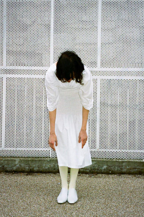 woman in a white dress, tights and shoes, standing before a tight white metal fence - bowed down towards the camera so we see the top of her head