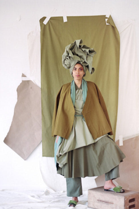 Woman in a multi-layerd outfit in greens and greys - in a photo studio with green fabric taped to the wall