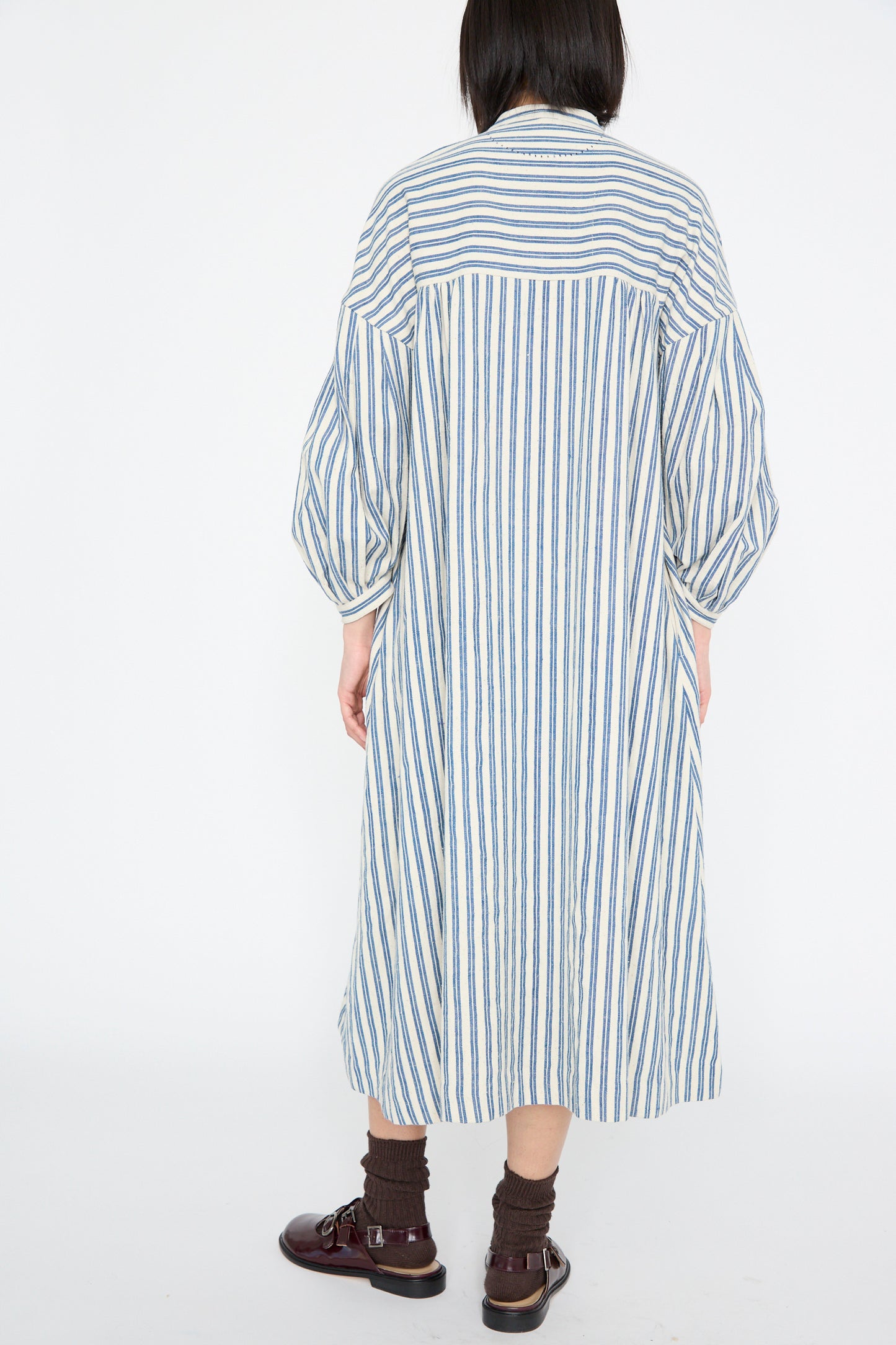 A person stands against a plain backdrop, wearing the 11.11 Cotton Gurue Dress in Medium Indigo Stripe with an A-line silhouette, and paired with dark shoes.
