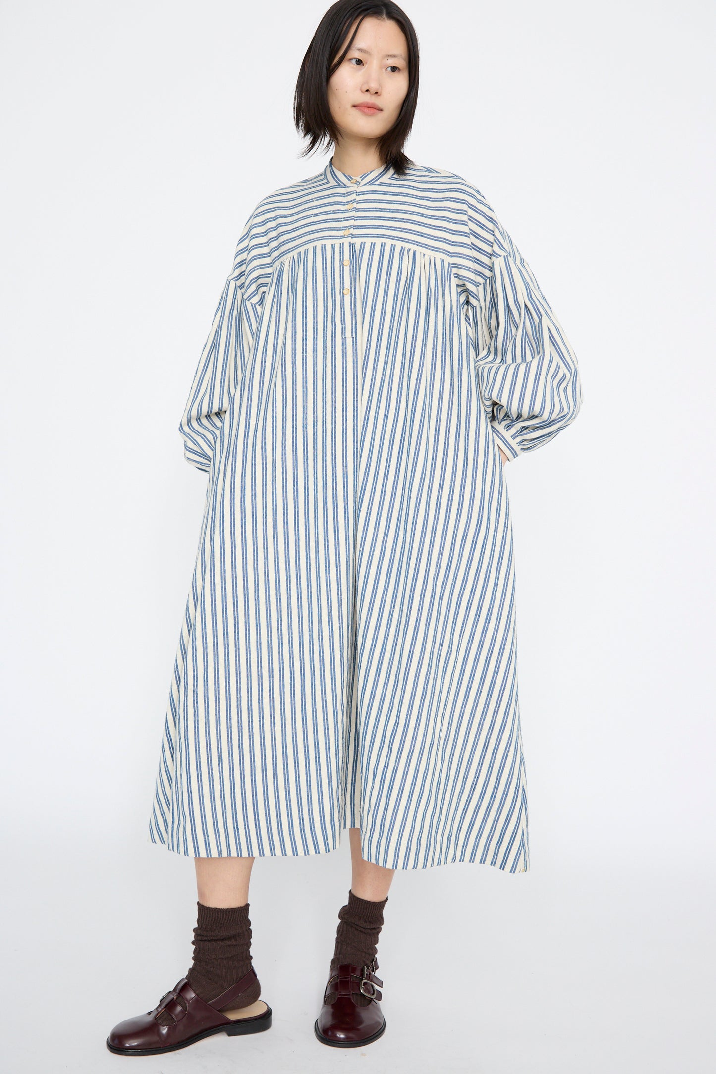A model wearing the 11.11 Cotton Gurue Dress in Medium Indigo Stripe, with brown shoes and socks stands against a plain white background. The dress has a loose, A-line silhouette and long sleeves made of handwoven cotton.