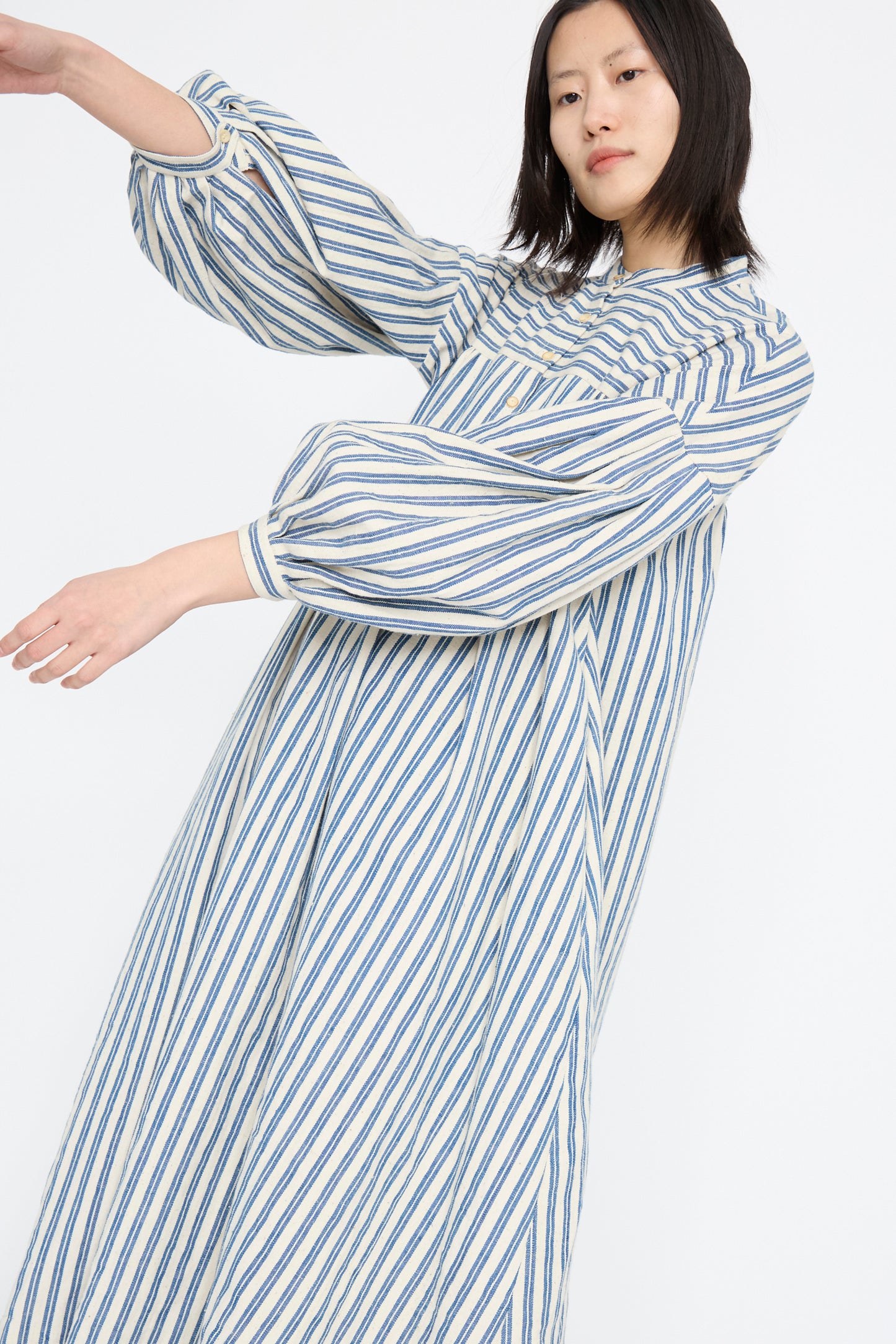 A person models the Cotton Gurue Dress by 11.11, featuring medium indigo stripes and billowy sleeves, posed against a plain background.