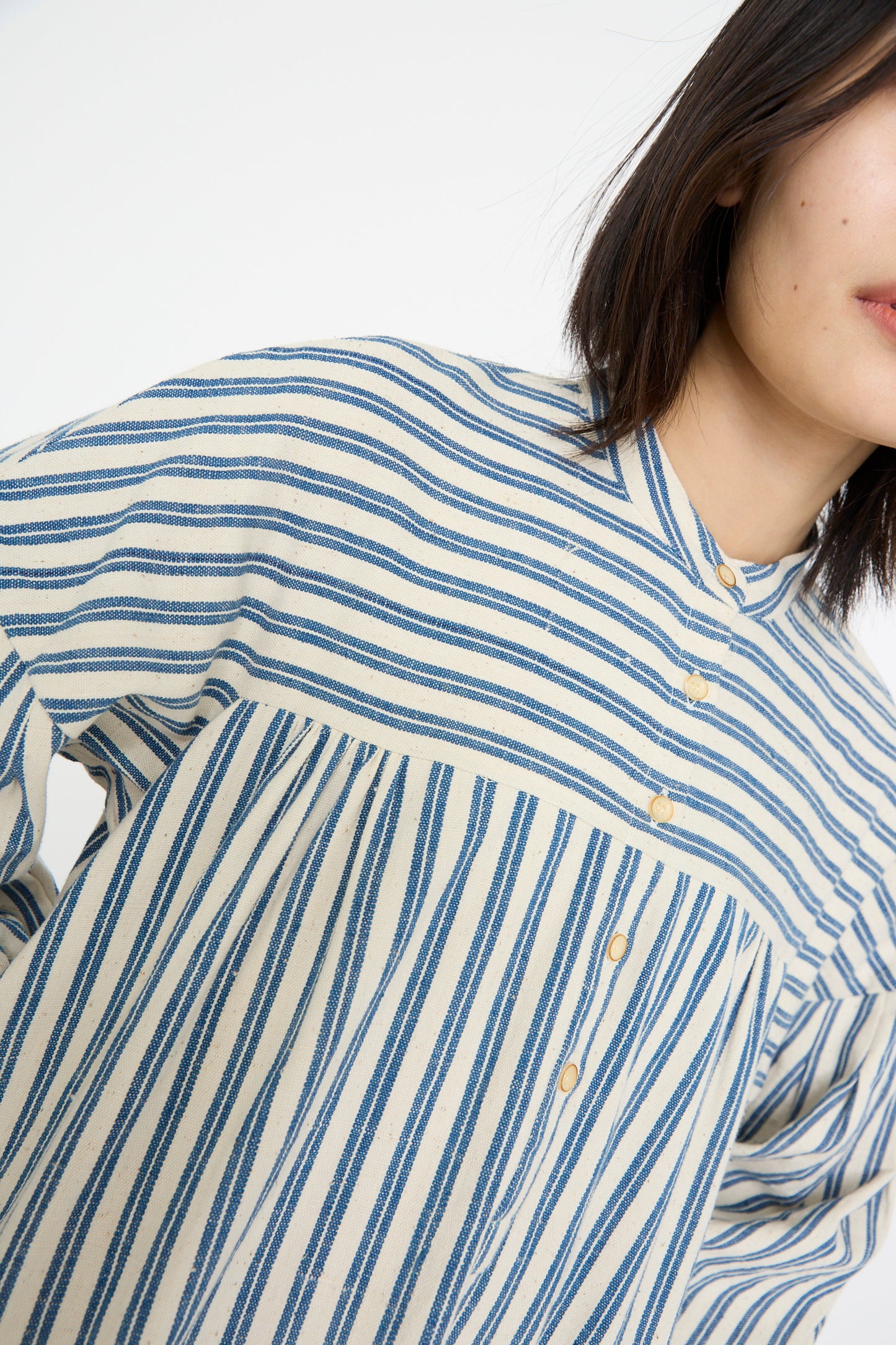 A model wearing the 11.11 Cotton Gurue Dress in Medium Indigo Stripe, made from handwoven cotton, is partially visible against a plain background, blurred from mid-torso up.