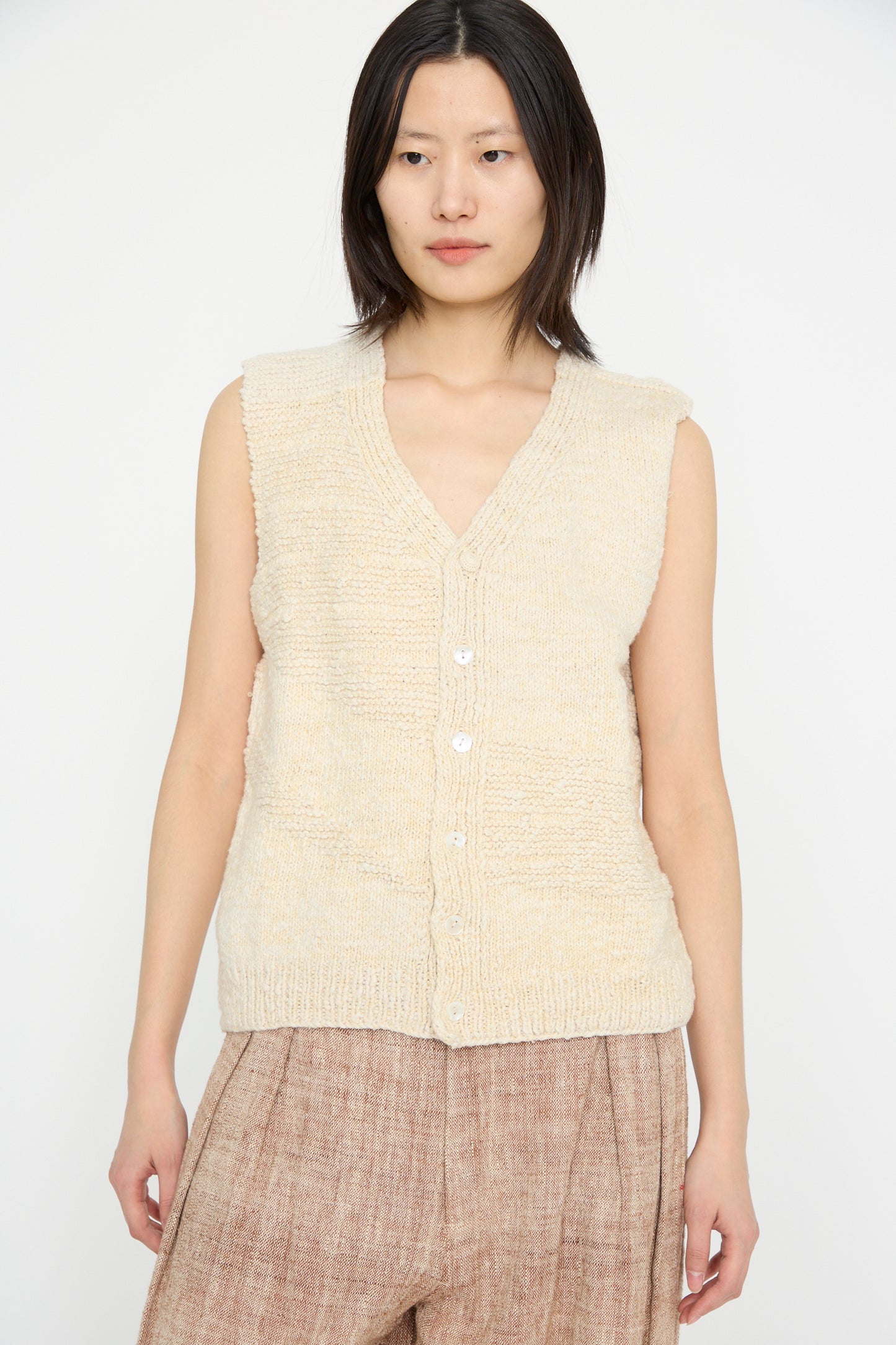 A person is wearing the Oro Vest in Ecru by 11.11, a beige sleeveless knit top made of Merino wool, paired with brown pants against a plain white background.