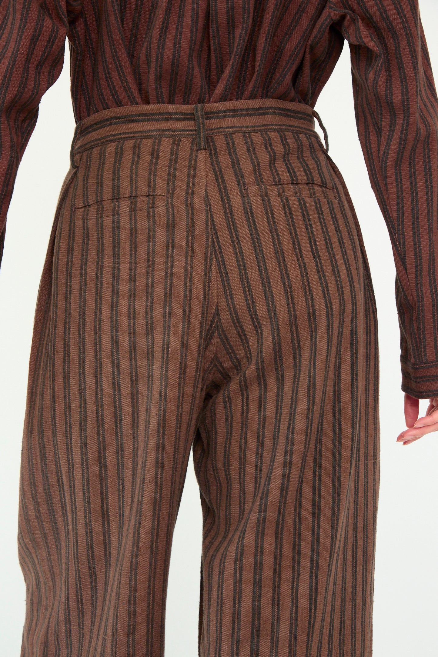 A person is seen from the back wearing 11.11's Tel Trouser in Burnt Umber Stripe, paired with a brown striped shirt, with one arm slightly visible, against a white backdrop.