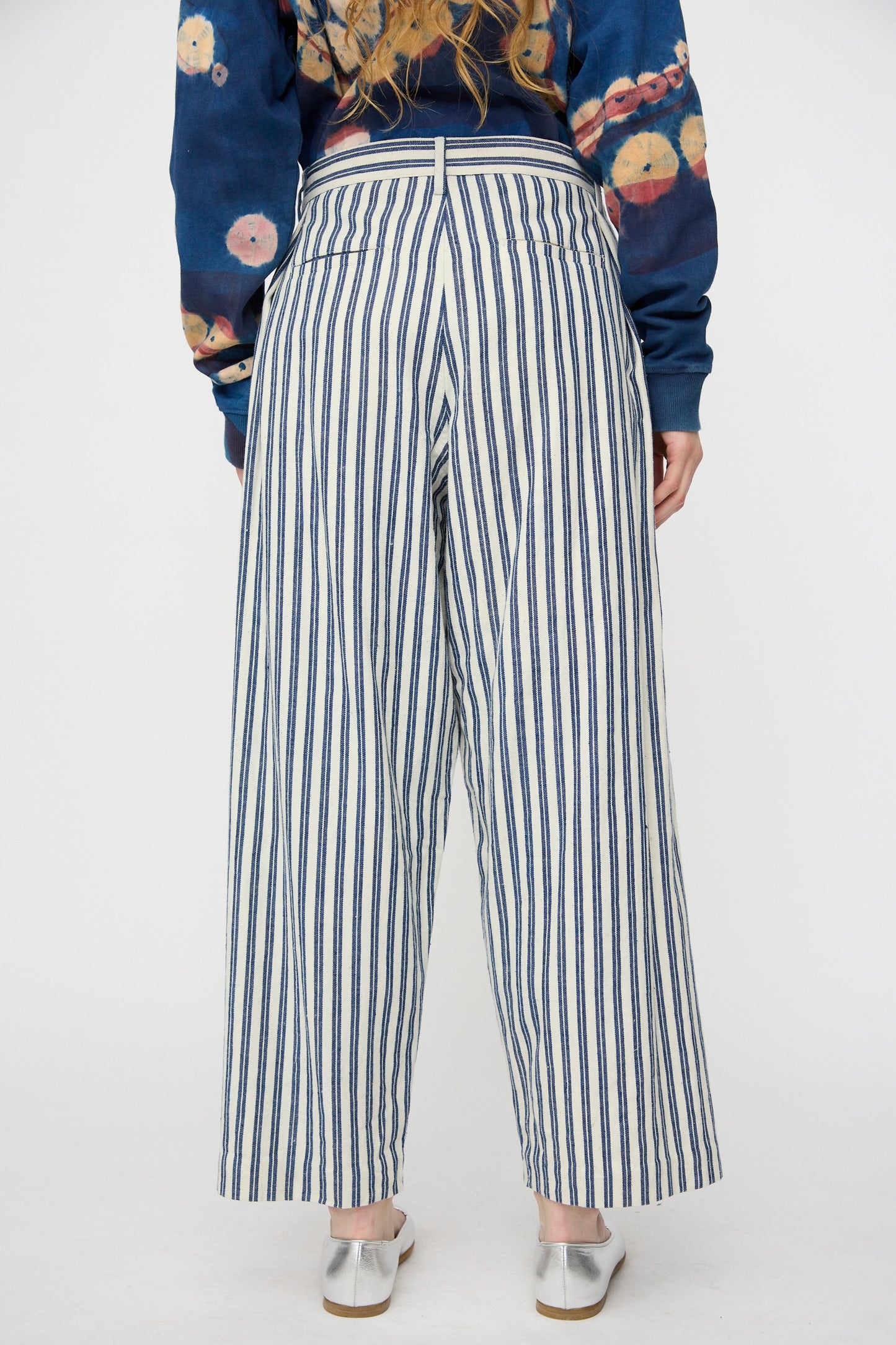 A person stands facing away against a plain background, wearing 11.11's Tel Trouser in Medium Indigo Stripe, a printed long-sleeve shirt, and white shoes. The high-waisted wide-leg trousers are crafted from organic cotton.