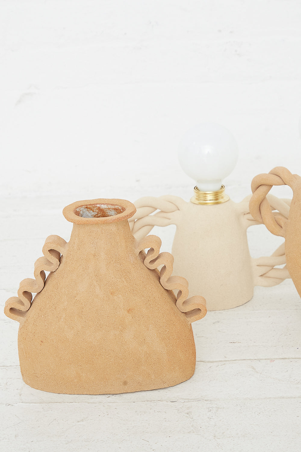 Two ceramic objects with unique shapes, one resembling an Amphora Soleil vase in Raw Sunny Brown Clay and the other a lamp, displayed against a white background by Clandestine.