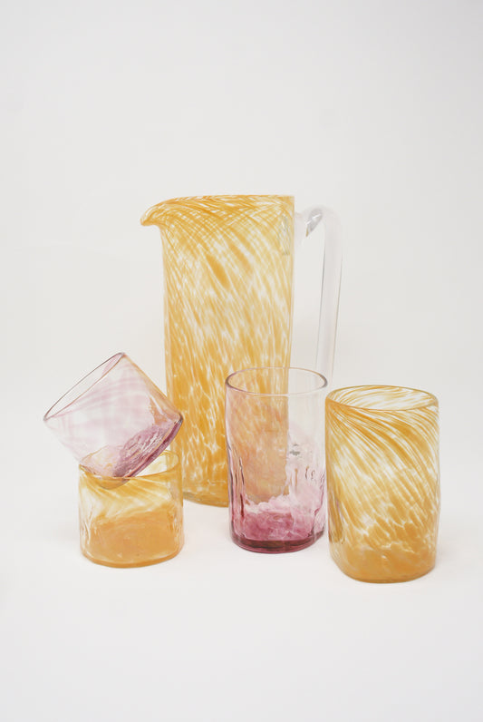 A set of Xaquixe handblown glassware with swirling yellow and pink patterns, including a Large Saffron Recycled Glass Pitcher and three different sized cups, made from recycled glass.