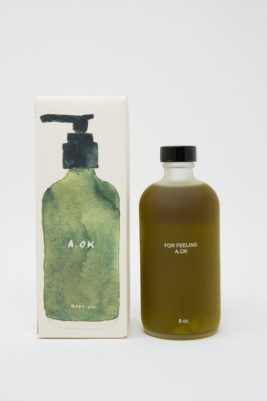 A bottle of Botanical Body Oil labeled "FOR FEELING A.OK" is positioned next to its packaging, which has an illustration of a green bottle with a pump and the text "A.OK Botanial Body Oil." Infused with essential oils, this 8 oz unisex aroma blend is perfect for everyone.