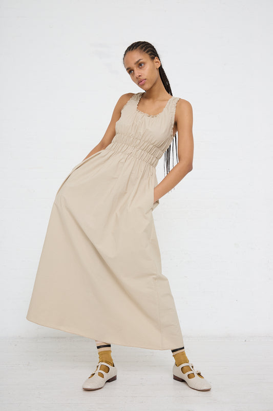 A woman poses in a AVN light beige sleeveless midi Accordion Dress made of lightweight cotton with a ruched bodice, paired with white sandals, against a white background.