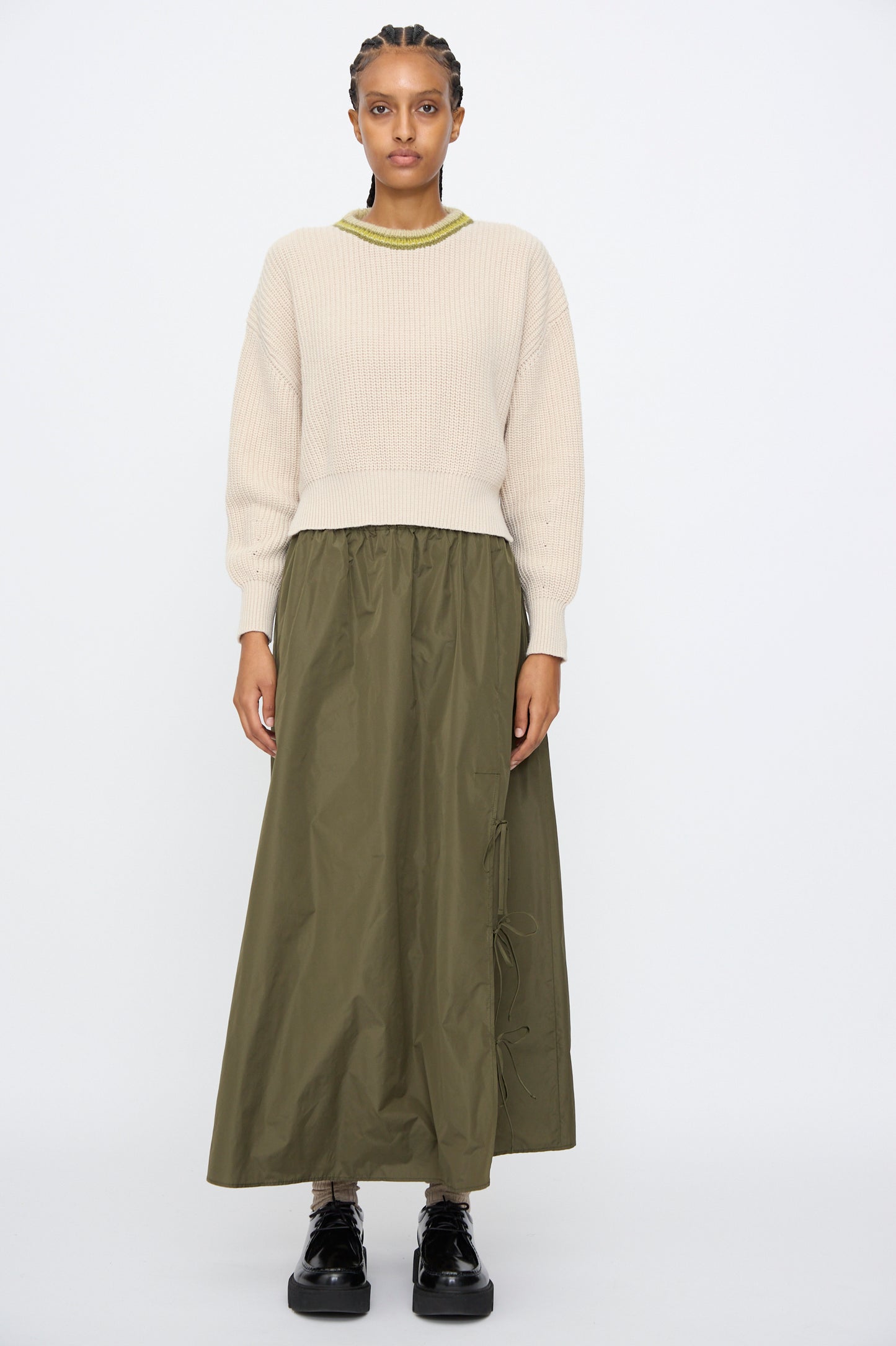 A person stands facing forward against a plain white background, wearing the AVN American Sweater in Sand, an olive green maxi skirt, and black platform shoes.