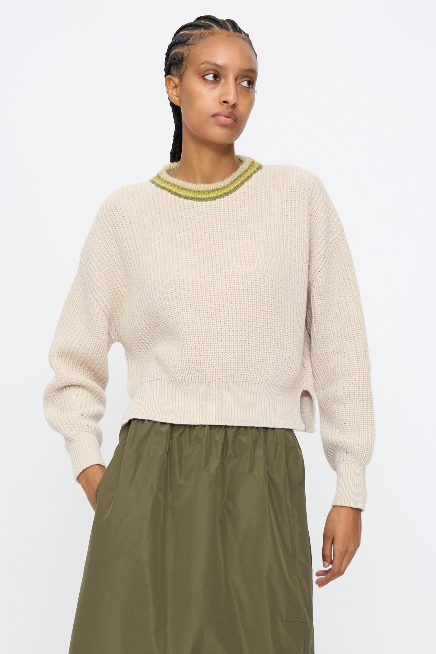 A woman wearing the AVN American Sweater in Sand, featuring a ribbed texture and green-trimmed neckline, pairs it with an olive-green skirt while standing against a plain white background.