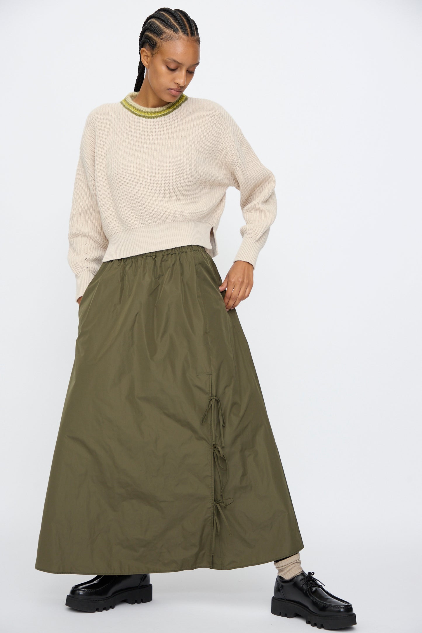A person wears the AVN American Sweater in Sand, paired with an olive green ruched maxi skirt, beige socks, and black shoes, standing against a plain white background.