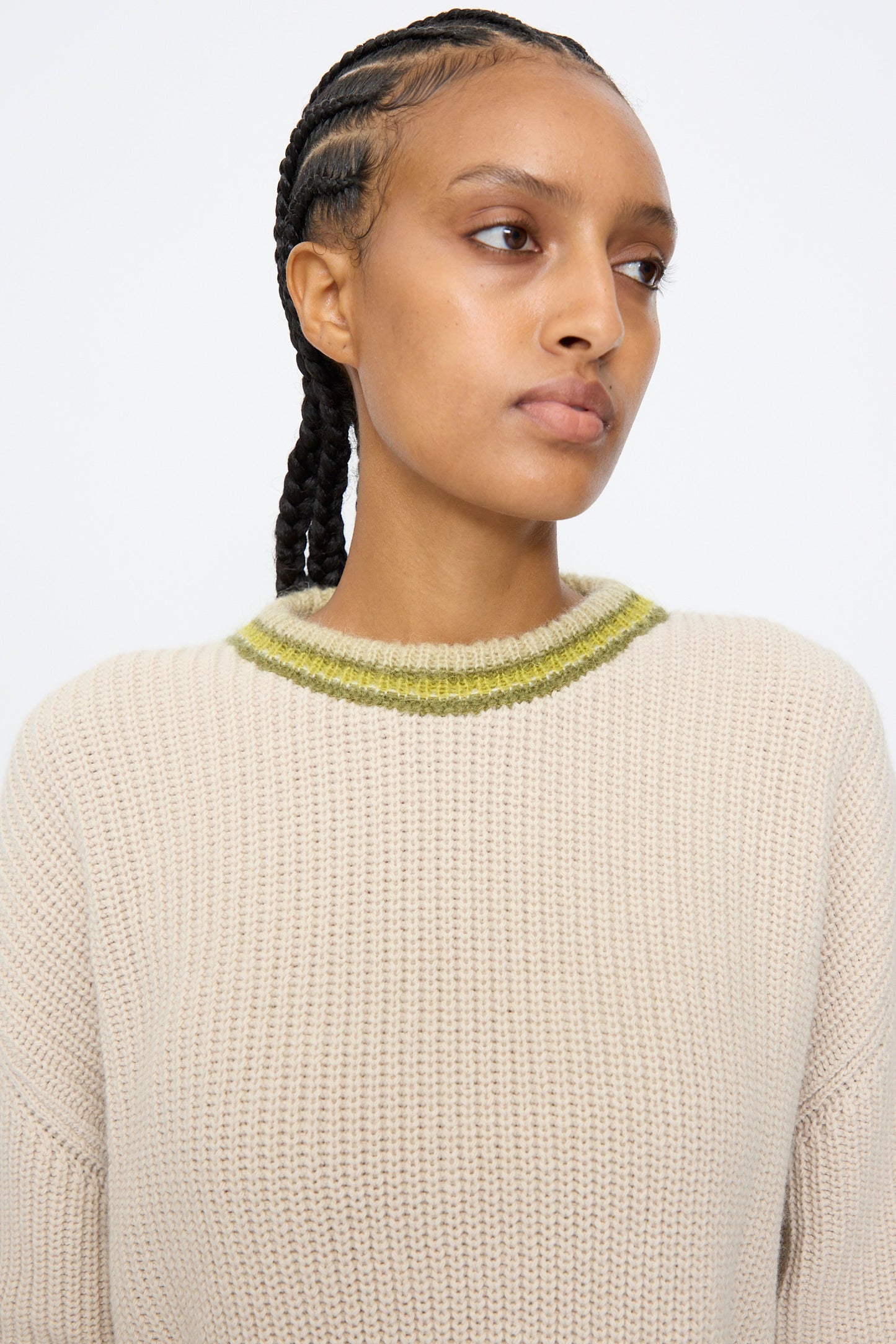 A person with braided hair wears the AVN American Sweater in Sand, featuring a green-striped collar, and looks to the side against a plain background.