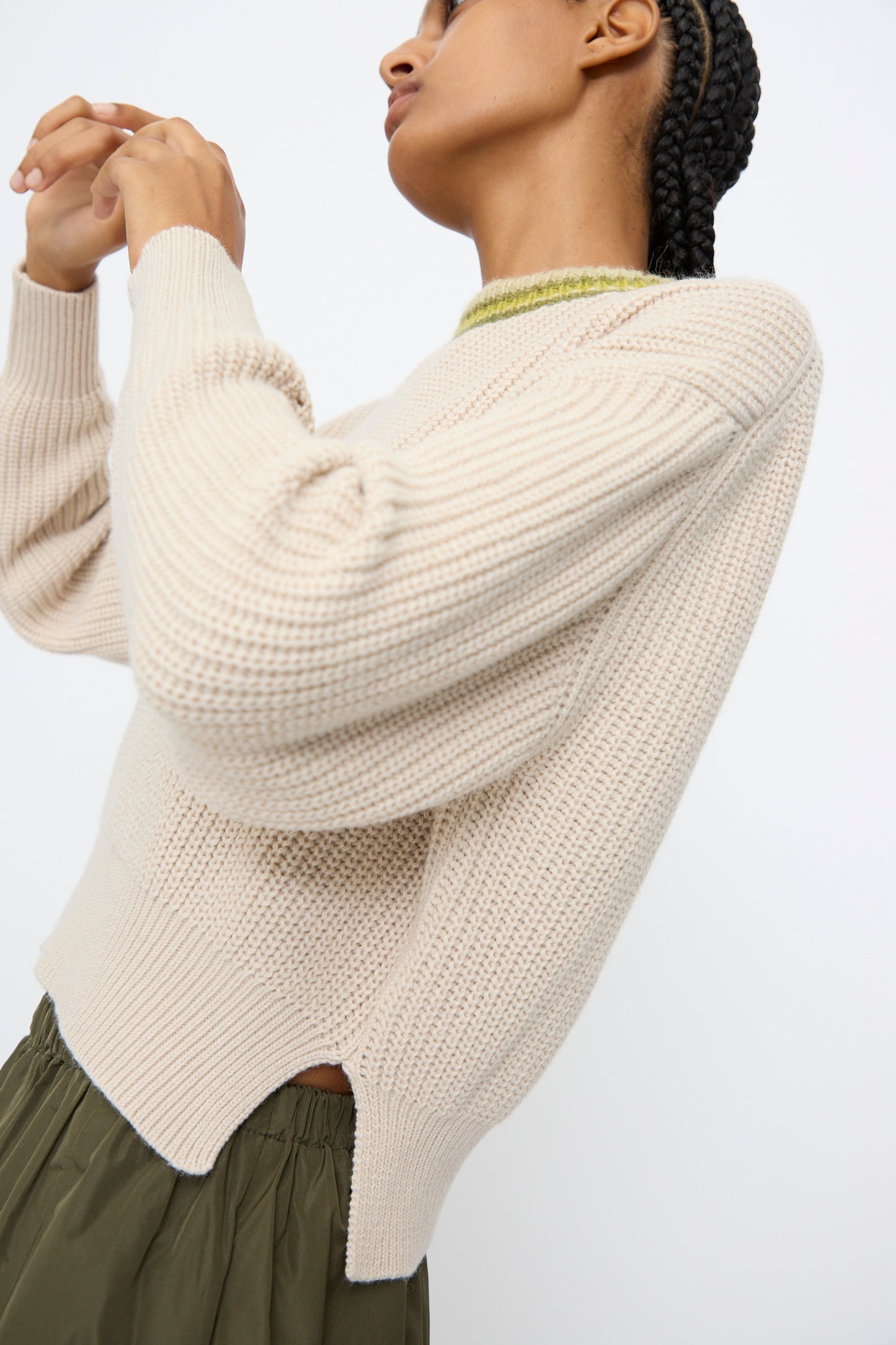 Person with braided hair wearing the AVN American Sweater in Sand—a cream ribbed wool piece with a green collar—paired with an olive skirt, posed against a plain background.