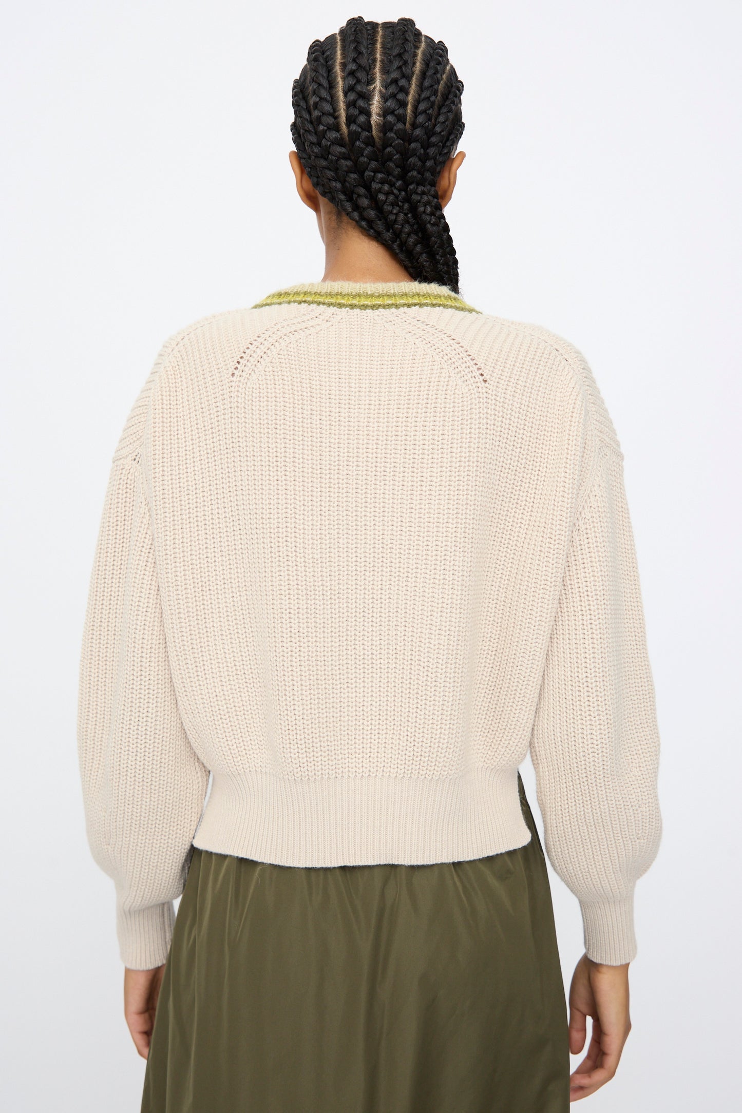 A person with braided hair is seen from the back, wearing the AVN American Sweater in Sand and an olive-green skirt.