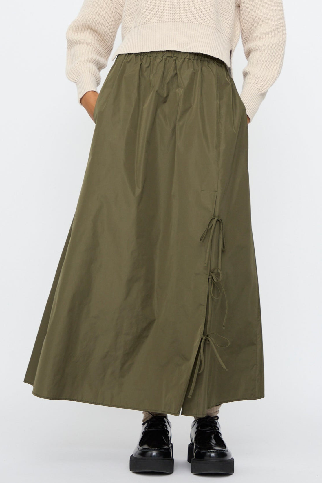 A person wears a cream ribbed sweater with the AVN Bows Skirt in Military Green, featuring an A-line silhouette and front tie details, paired with black platform shoes.