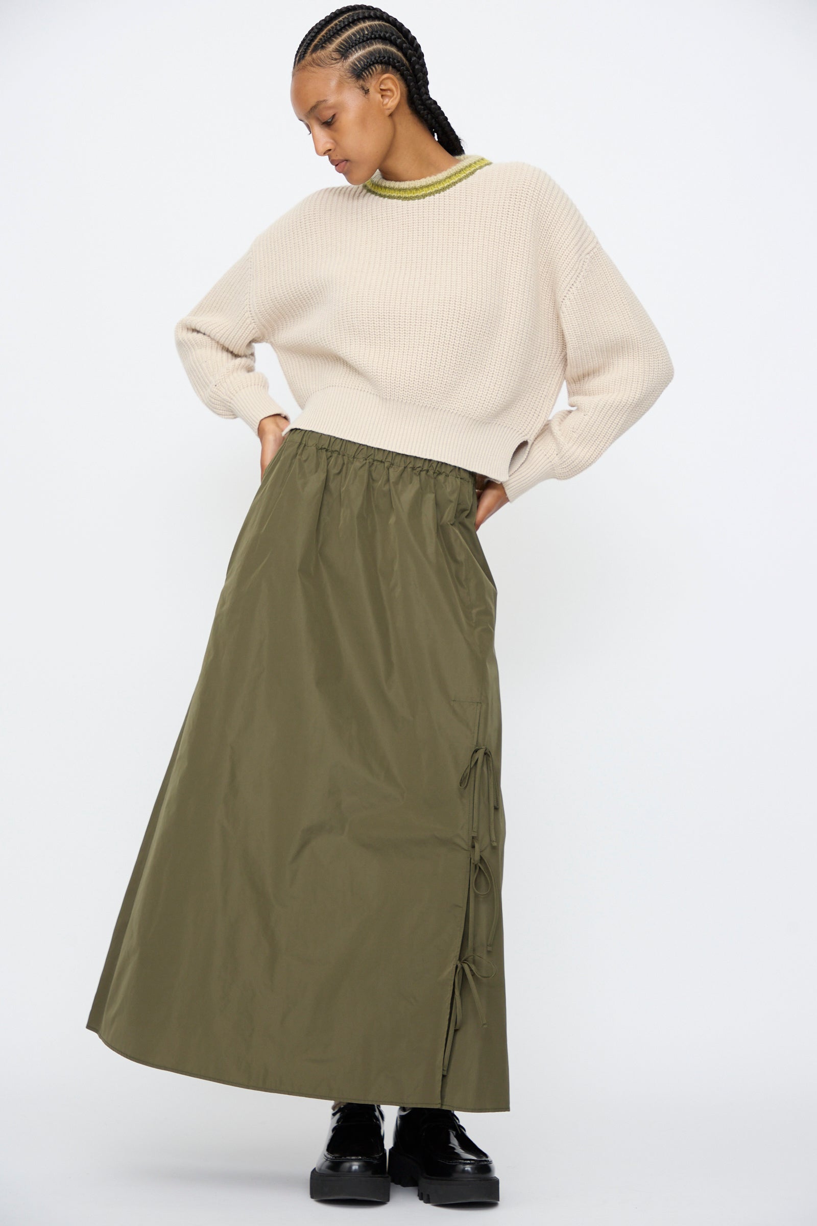 A person wears a cream knit sweater with AVN's Bows Skirt in Military Green, featuring an A-line silhouette and side ties, paired with black platform shoes, standing against a plain white background.