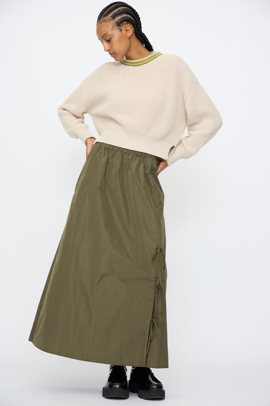 A person wears a cream knit sweater with AVN's Bows Skirt in Military Green, featuring an A-line silhouette and side ties, paired with black platform shoes, standing against a plain white background.