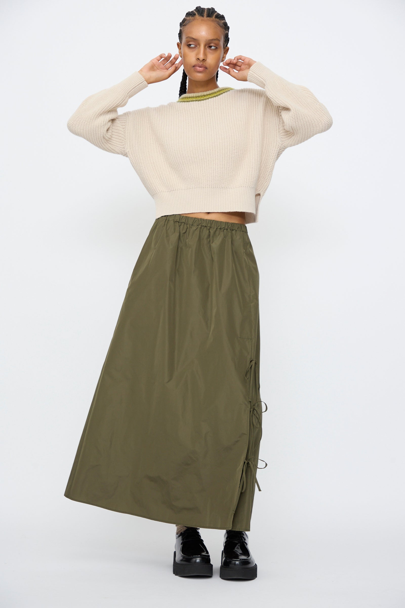 A person stands against a plain background wearing a cream cropped sweater, AVN's Bows Skirt in Military Green with side ties and an A-line silhouette, paired with black platform shoes.