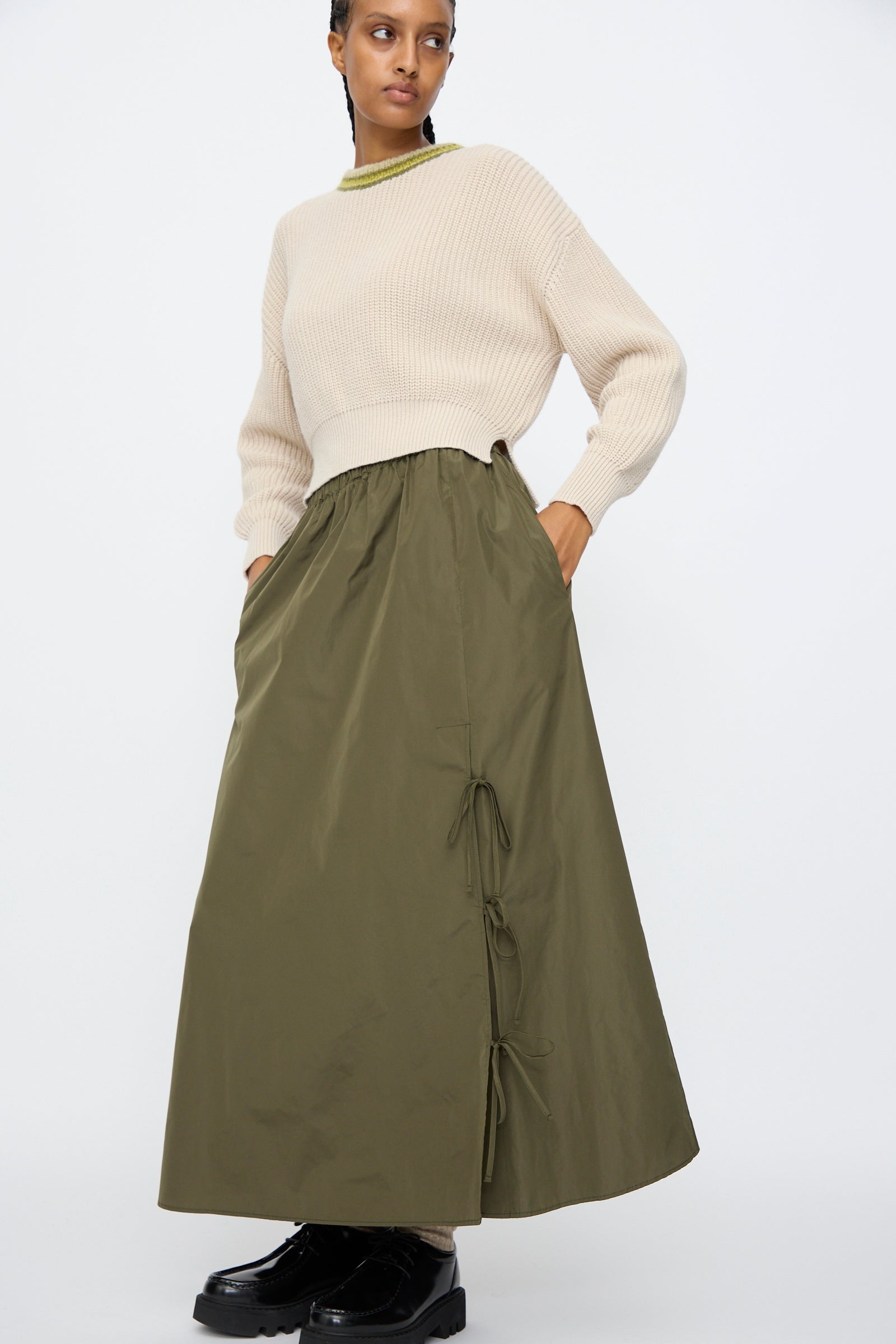 A person wears the AVN Bows Skirt in Military Green, featuring side ties and a soft A-line silhouette, paired with a cream knit sweater, standing with hands in pockets against a plain background.