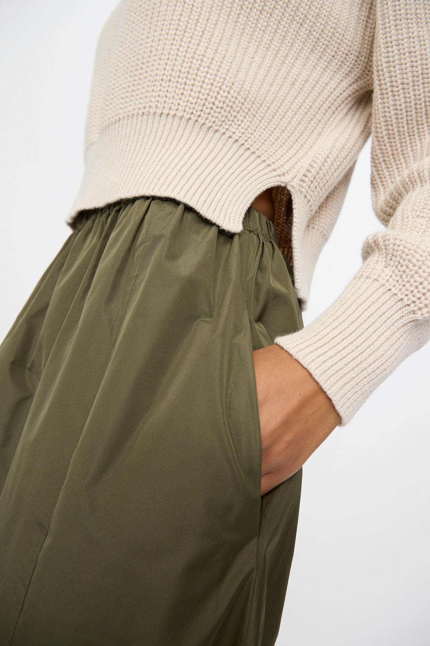 A person wears AVN's Bows Skirt in Military Green, styled with an off-white knitted sweater and their hand in the skirt pocket, highlighting the relaxed A-line silhouette.