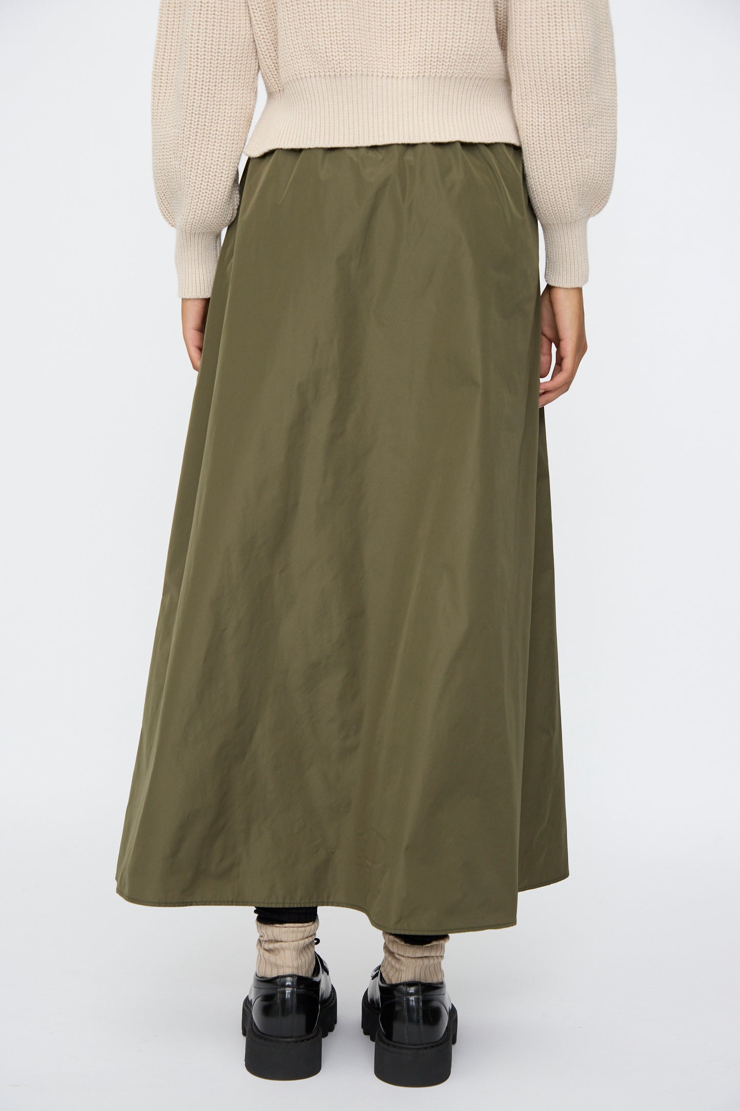 A person is seen from the back wearing AVN’s Bows Skirt in Military Green, paired with a beige sweater, black shoes, and light socks, set against a plain backdrop.
