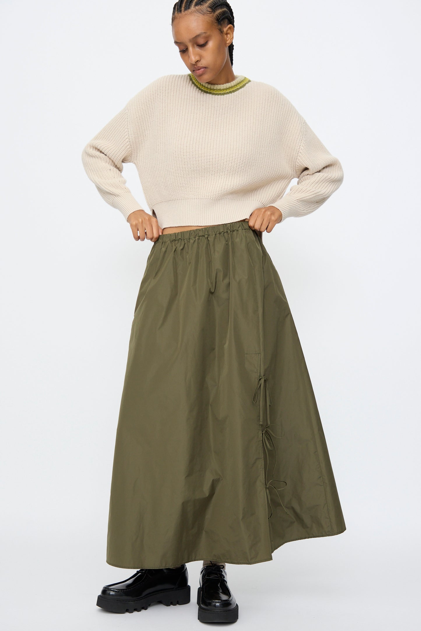 Model wears the AVN Bows Skirt in Military Green with drawstring details and an A-line silhouette, beige sweater, and black chunky shoes against a plain white background.