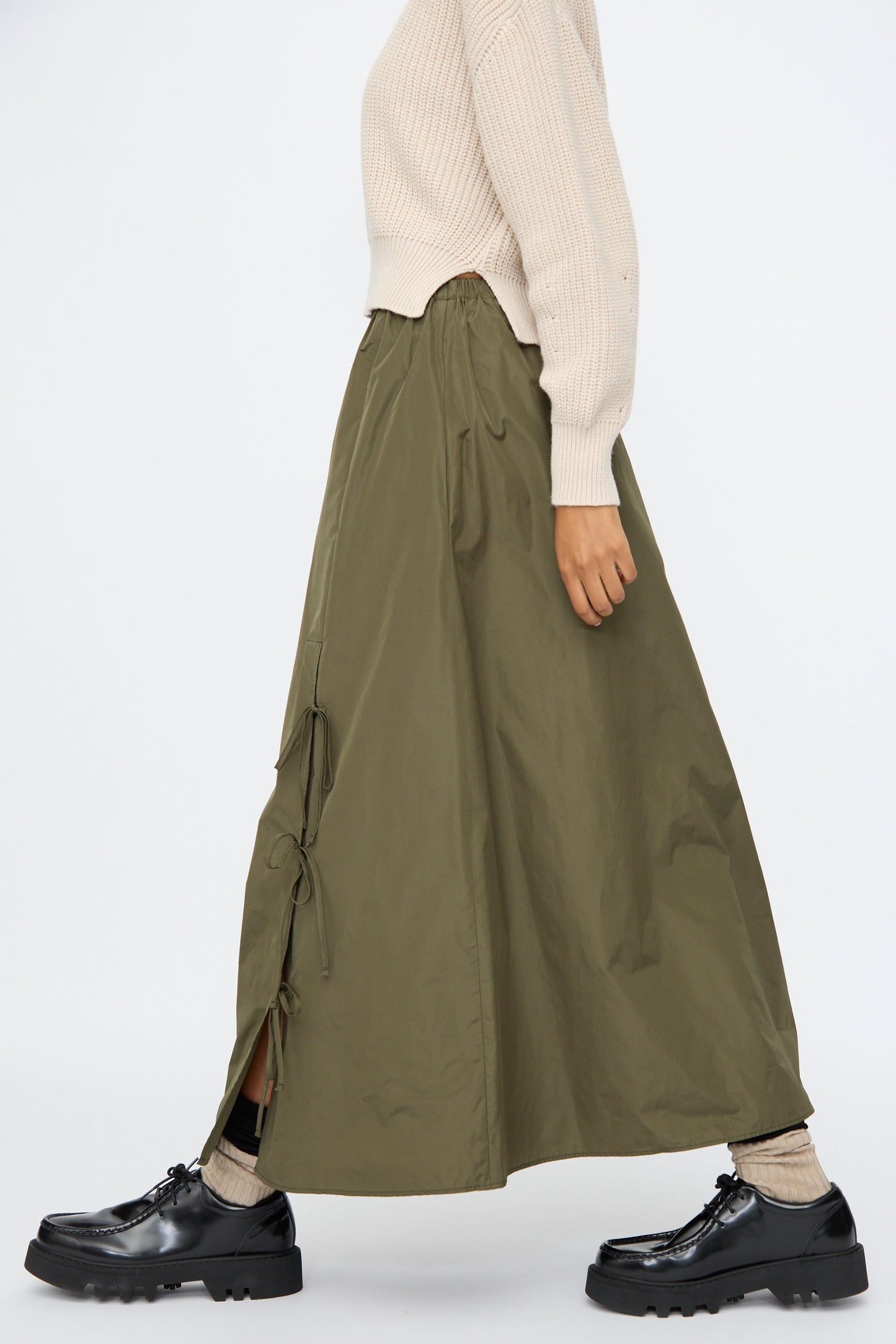 A person wears AVN's Bows Skirt in Military Green—a taffeta, A-line maxi with side ties—paired with a cream sweater, beige socks, and black chunky shoes, shown in profile against a plain background.