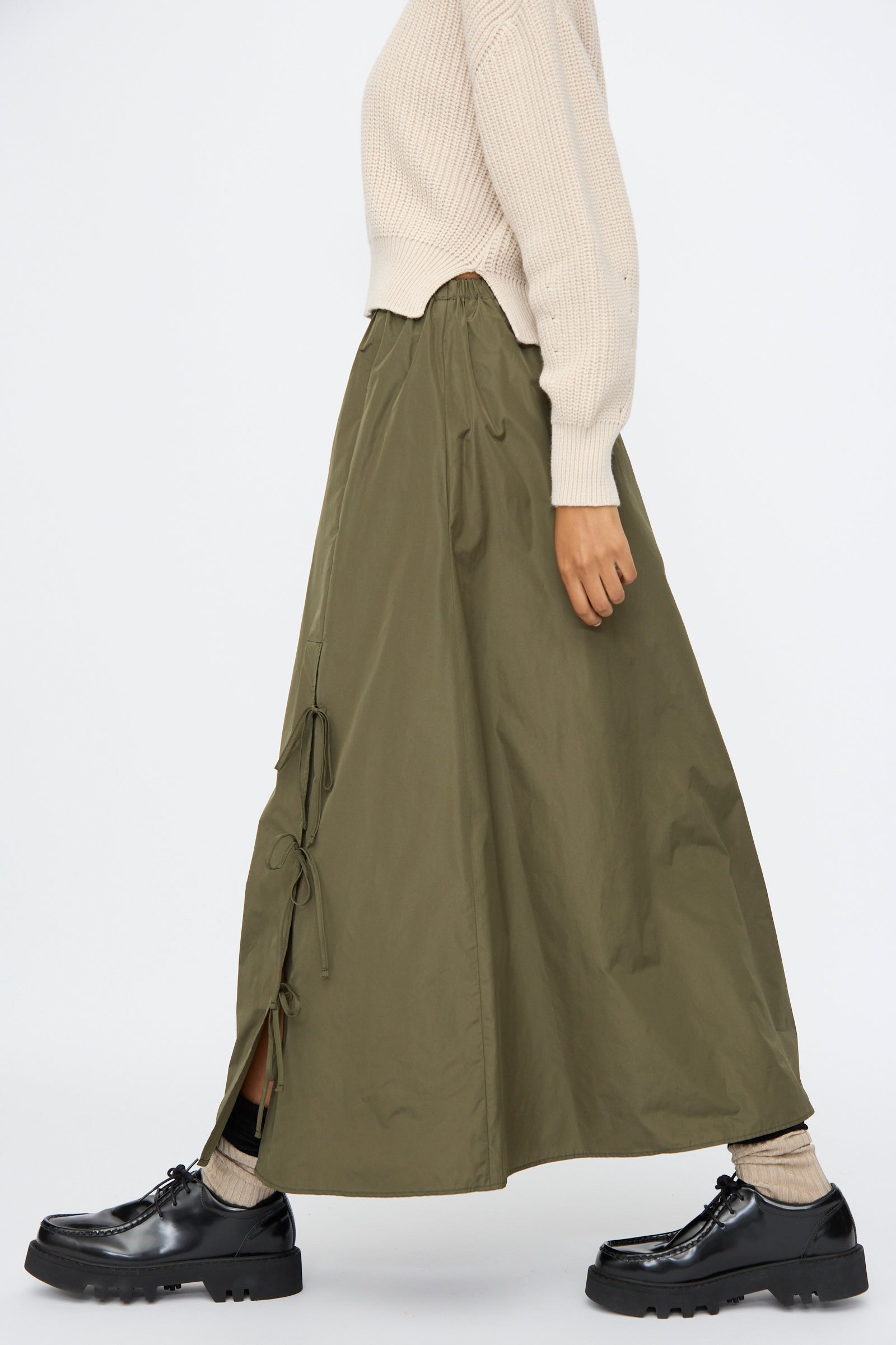 A person wears AVN's Bows Skirt in Military Green—a taffeta, A-line maxi with side ties—paired with a cream sweater, beige socks, and black chunky shoes, shown in profile against a plain background.