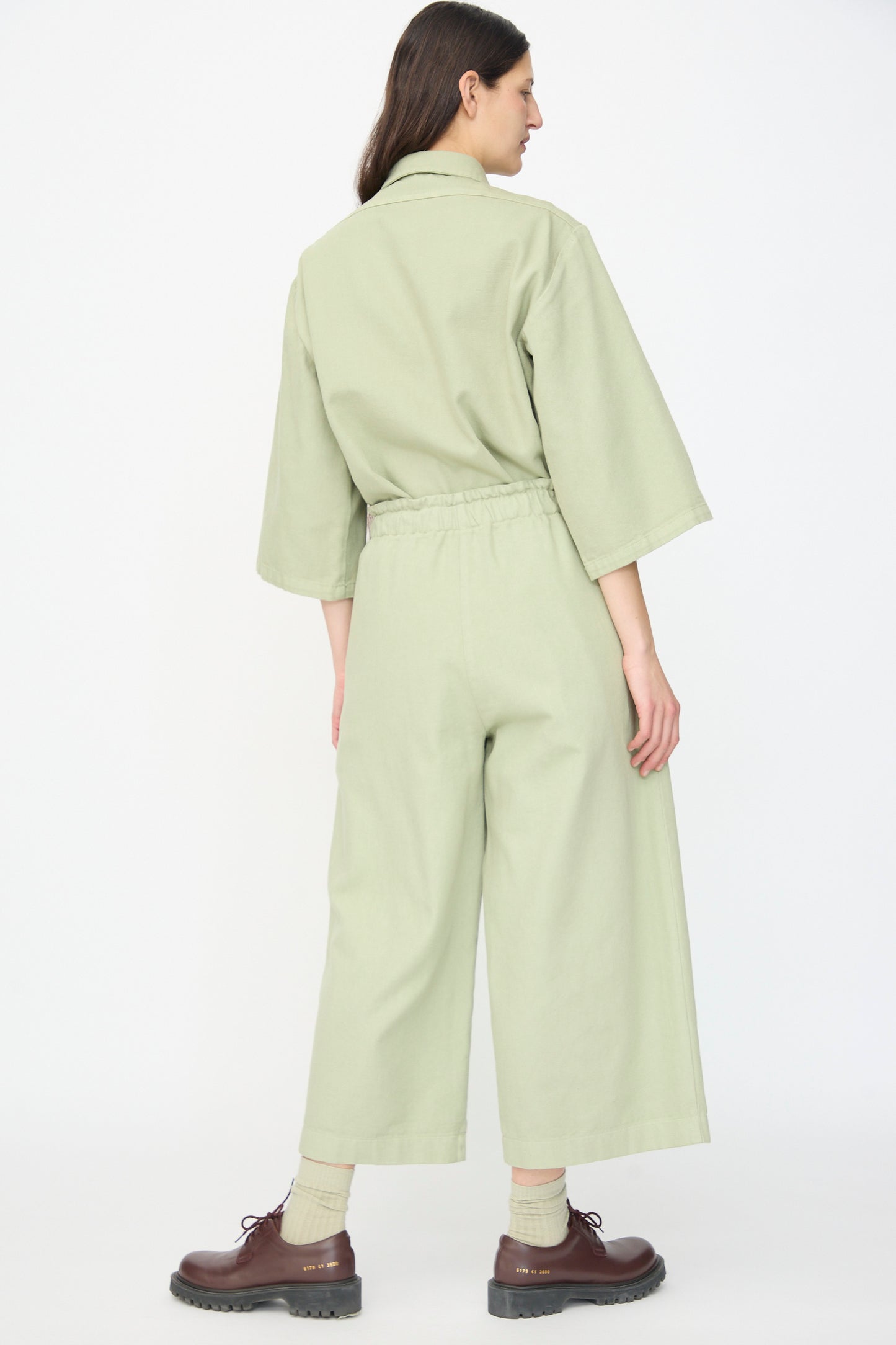 A person stands facing away, wearing AVN’s Easy Pants in Sage—wide-leg cotton trousers paired with brown shoes and light socks—set against a plain white background.