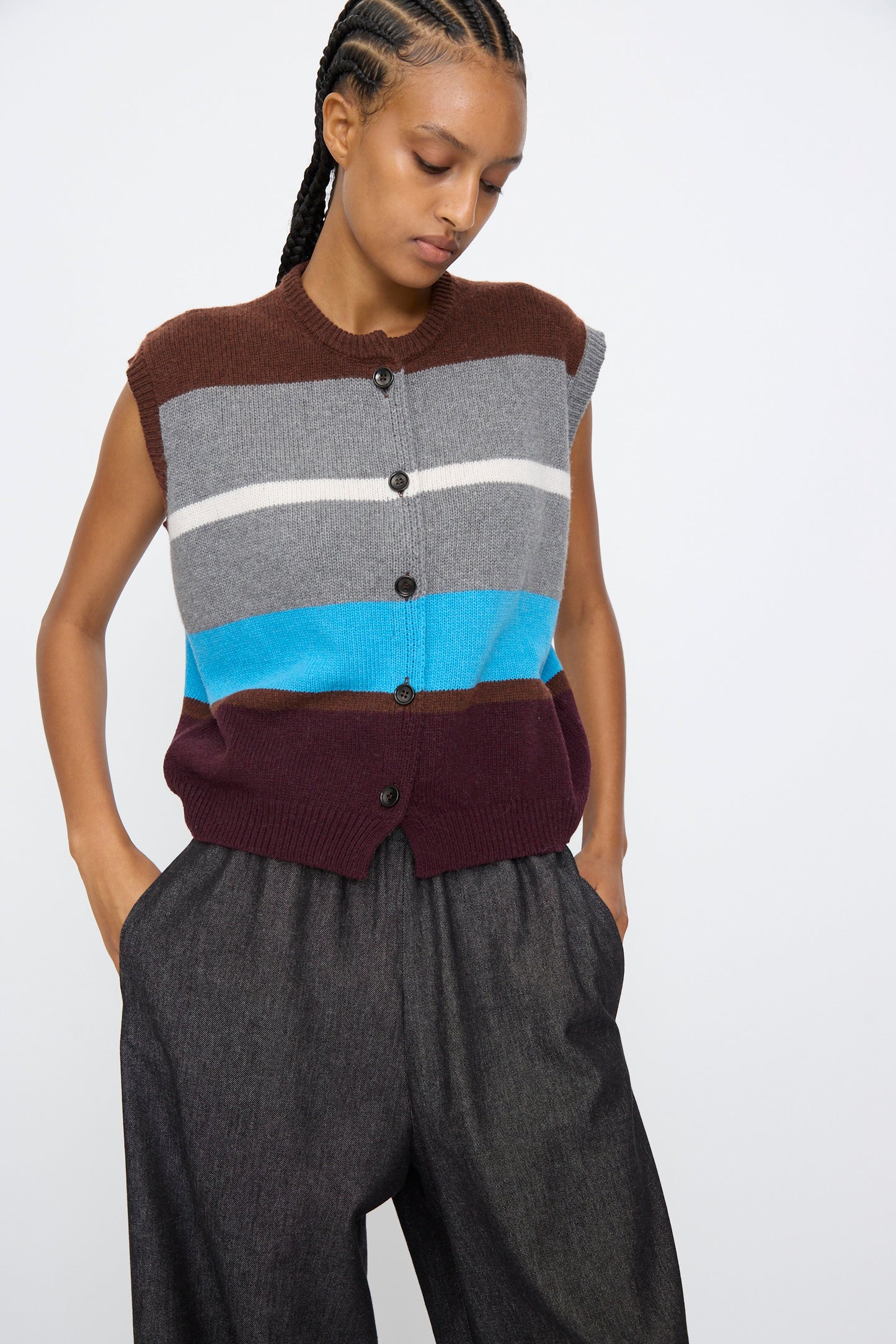 Wearing AVN’s Funky Vest in Multicolor—a sleeveless, button-up wool blend with brown, gray, white, blue, and burgundy stripes—paired with loose gray pants.