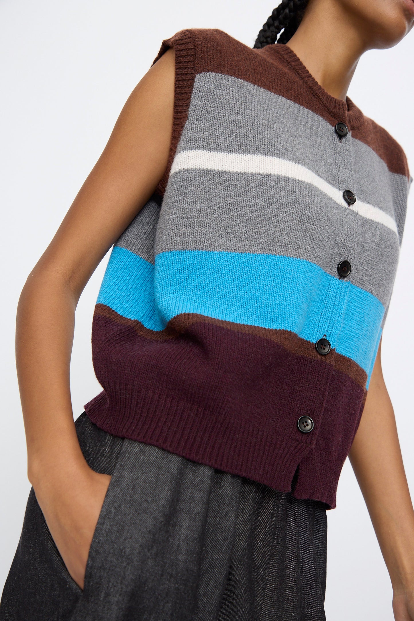 A person wears the AVN Funky Vest in Multicolor, a sleeveless wool-blend piece with brown, gray, white, blue, and burgundy stripes made in Italy, paired with dark gray high-waisted trousers.