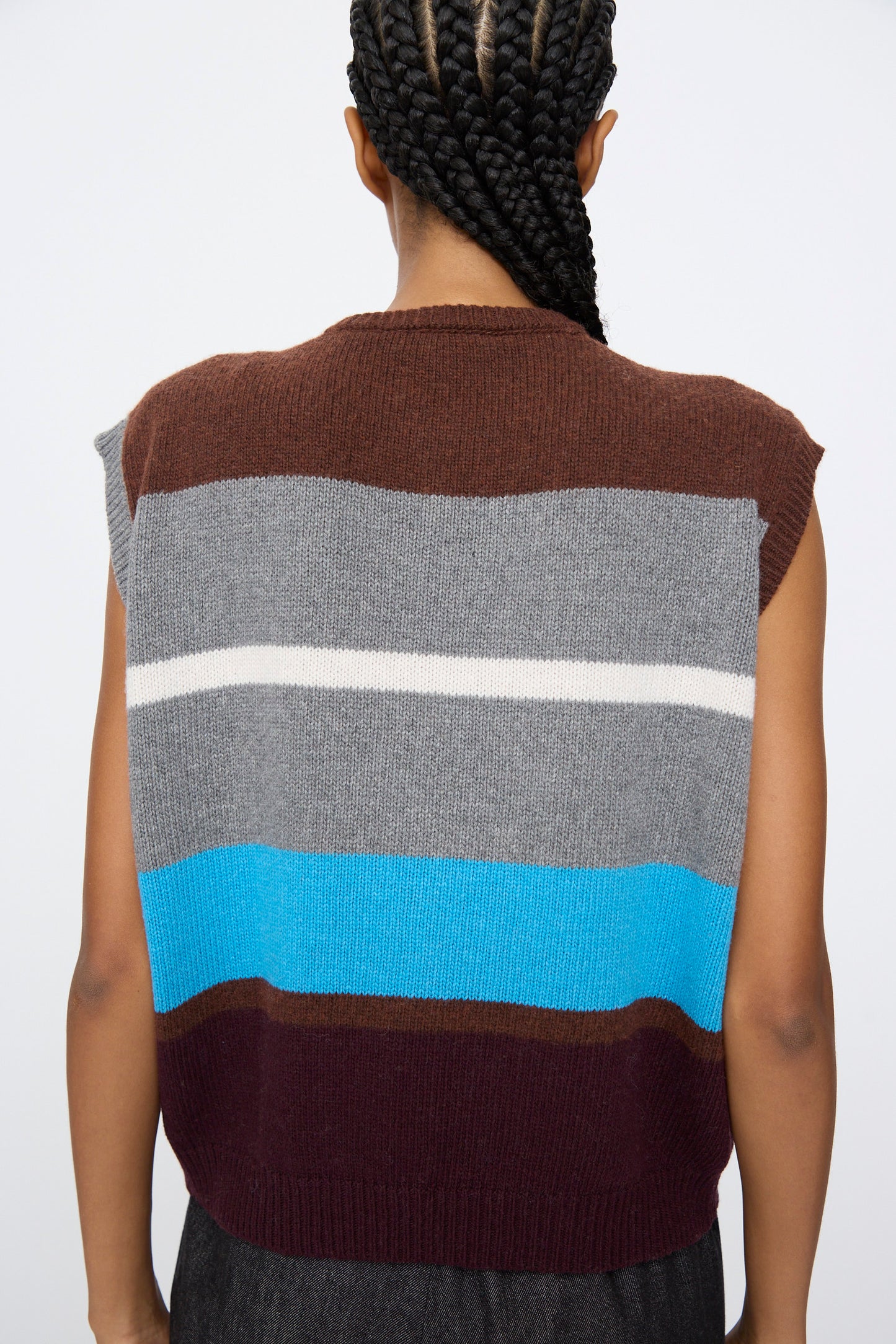 Person with braided hair, seen from behind, wears the AVN Funky Vest in Multicolor—a sleeveless wool blend vest featuring horizontal stripes in brown, gray, white, blue, and burgundy.