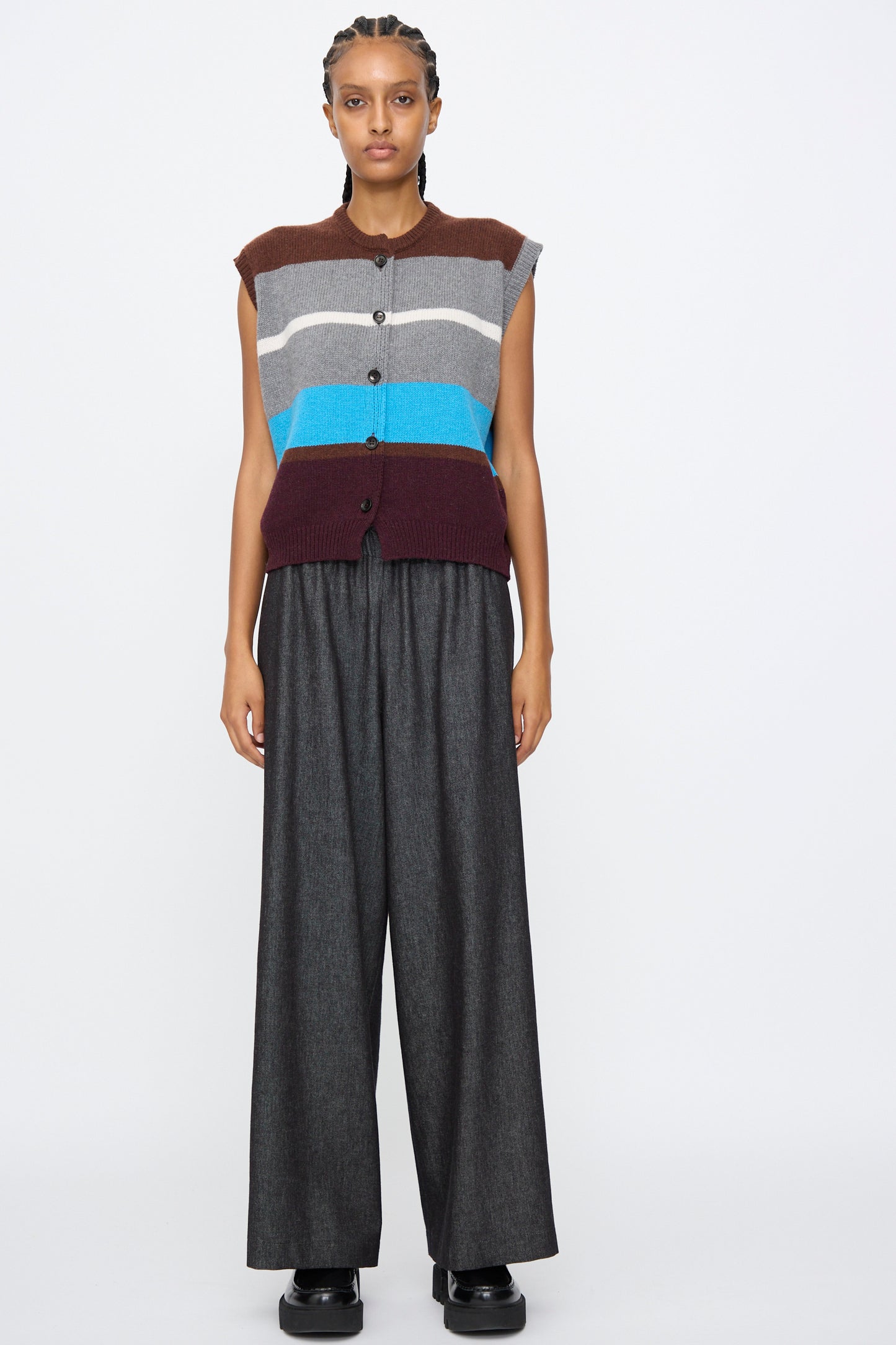 Person stands against a plain background in AVN's Long Easy Pants in Navy, featuring a wide leg and elasticated waistband, paired with a sleeveless striped button-up top and black shoes.