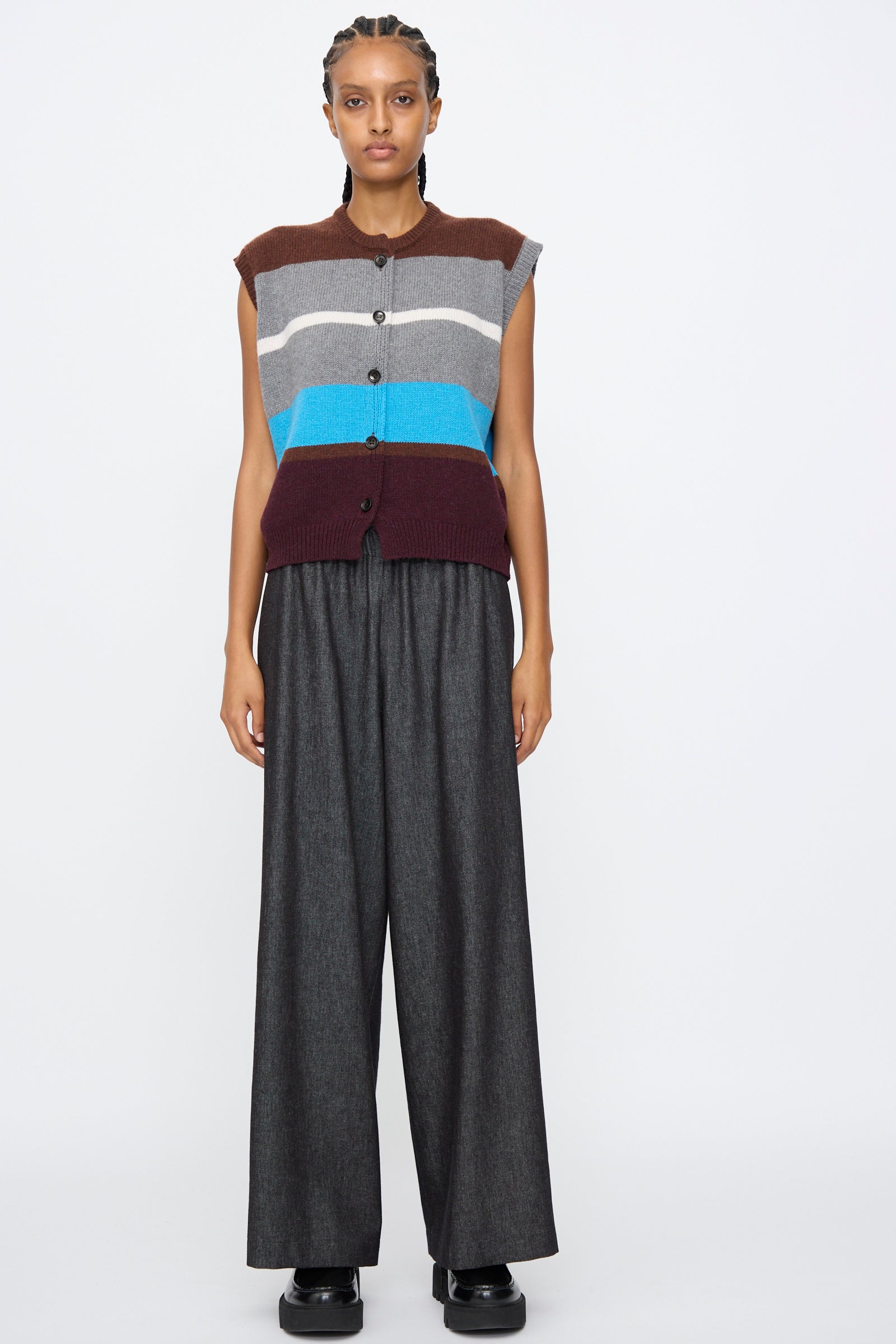 Person stands against a plain background in AVN's Long Easy Pants in Navy, featuring a wide leg and elasticated waistband, paired with a sleeveless striped button-up top and black shoes.