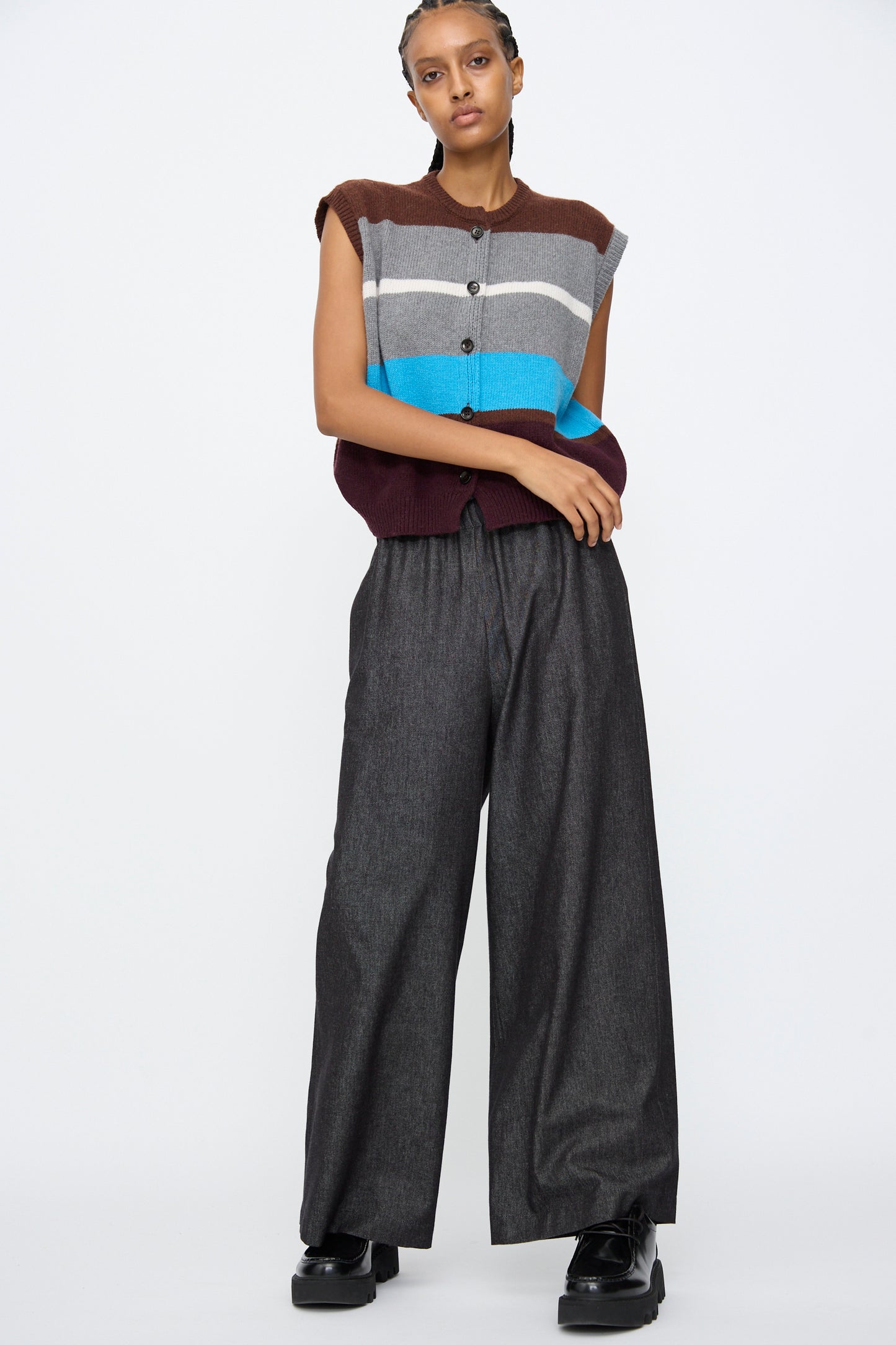 A person stands against a plain background wearing a sleeveless, striped sweater vest in brown, gray, blue, and maroon, paired with AVN's Long Easy Pants in Navy and black platform shoes.