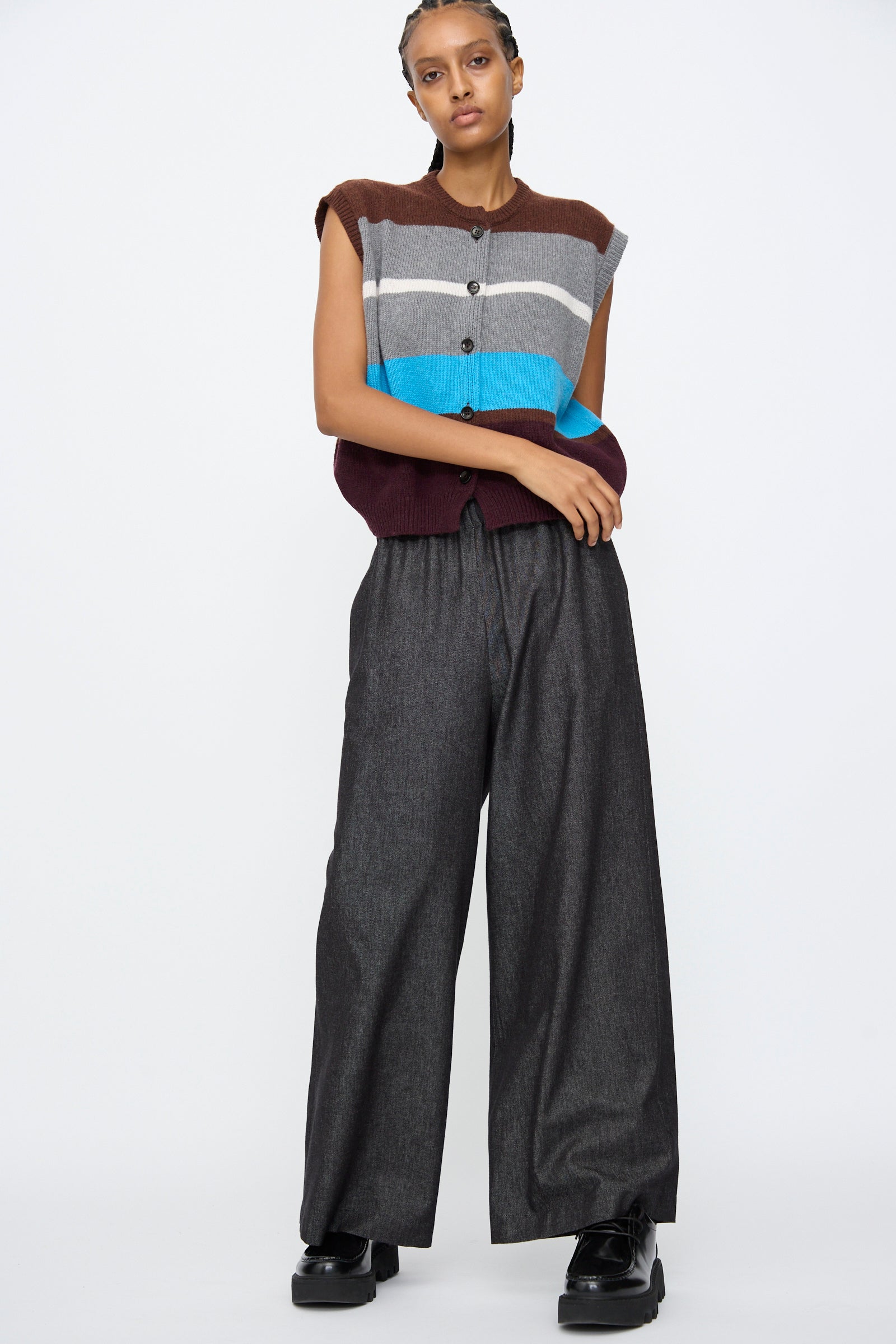 A person stands against a plain background wearing a sleeveless, striped sweater vest in brown, gray, blue, and maroon, paired with AVN's Long Easy Pants in Navy and black platform shoes.