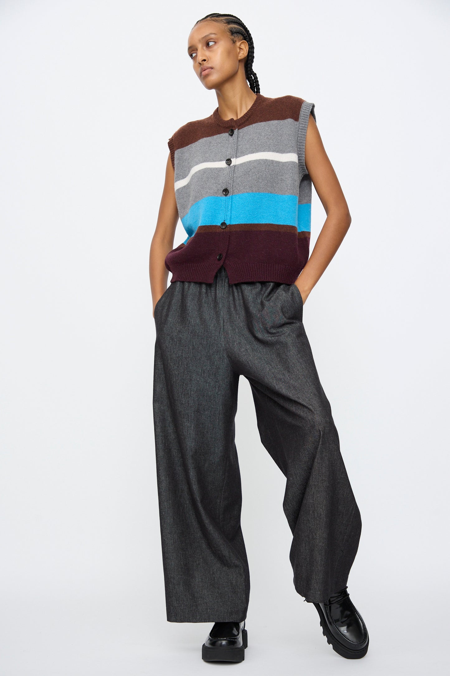 A person stands with hands in pockets wearing AVN's Long Easy Pants in Navy, styled with a sleeveless striped button-up sweater vest and black shoes, set against a plain white background.