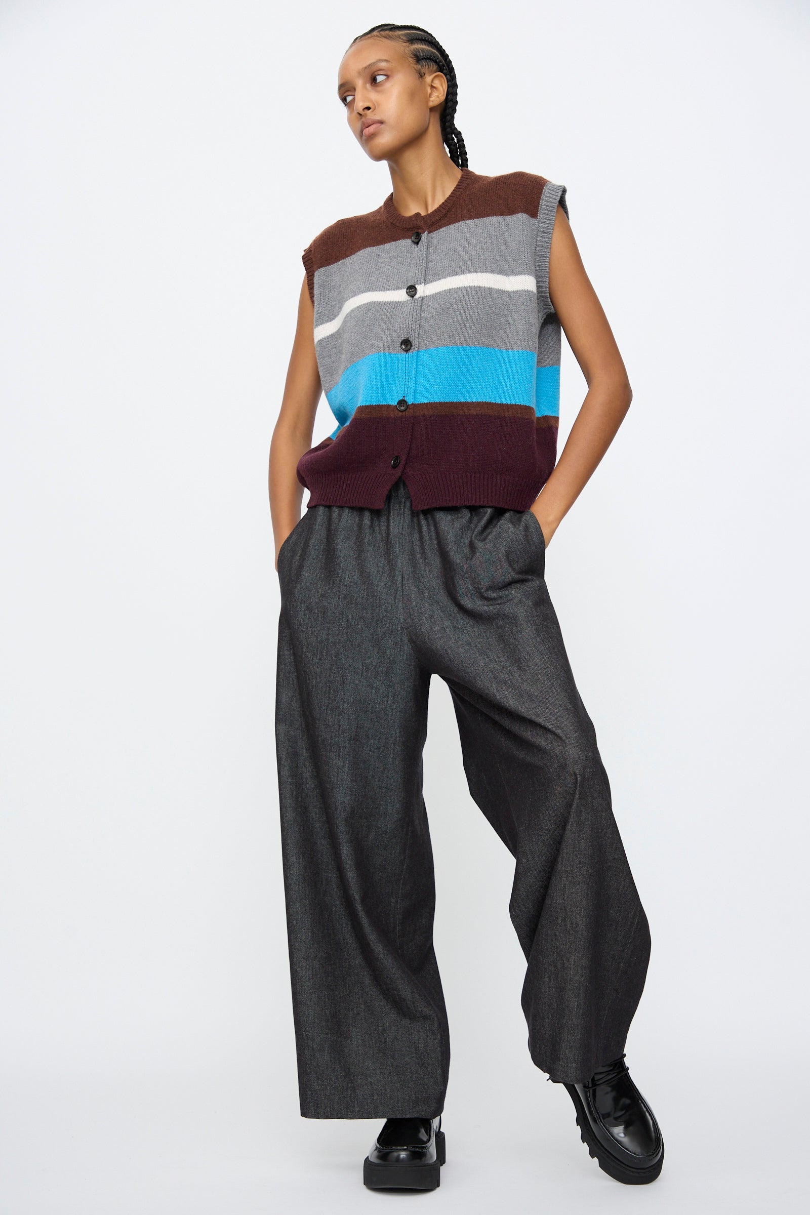 A person stands with hands in pockets wearing AVN's Long Easy Pants in Navy, styled with a sleeveless striped button-up sweater vest and black shoes, set against a plain white background.