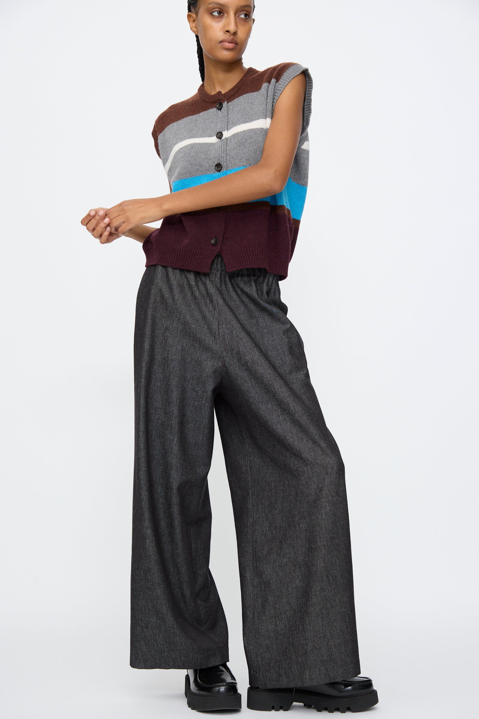 A person wears a sleeveless striped sweater vest, AVN Long Easy Pants in Navy with an elasticated waistband, and black shoes, standing against a plain white background.