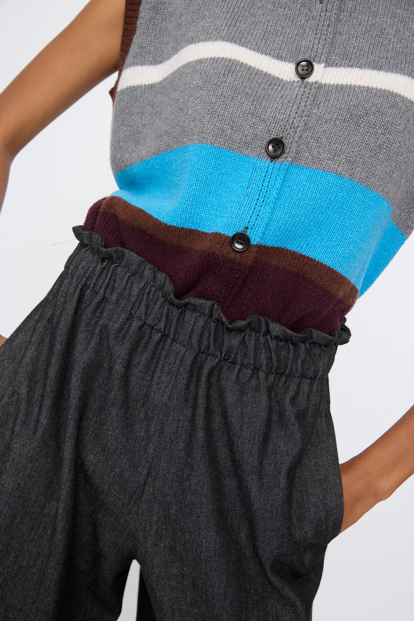 A person wears AVN’s Long Easy Pants in Navy—high-waisted, wide-leg with an elastic waistband—paired with a sleeveless, buttoned knit top in gray, blue, brown, and white stripes. Hands are tucked into the pockets.