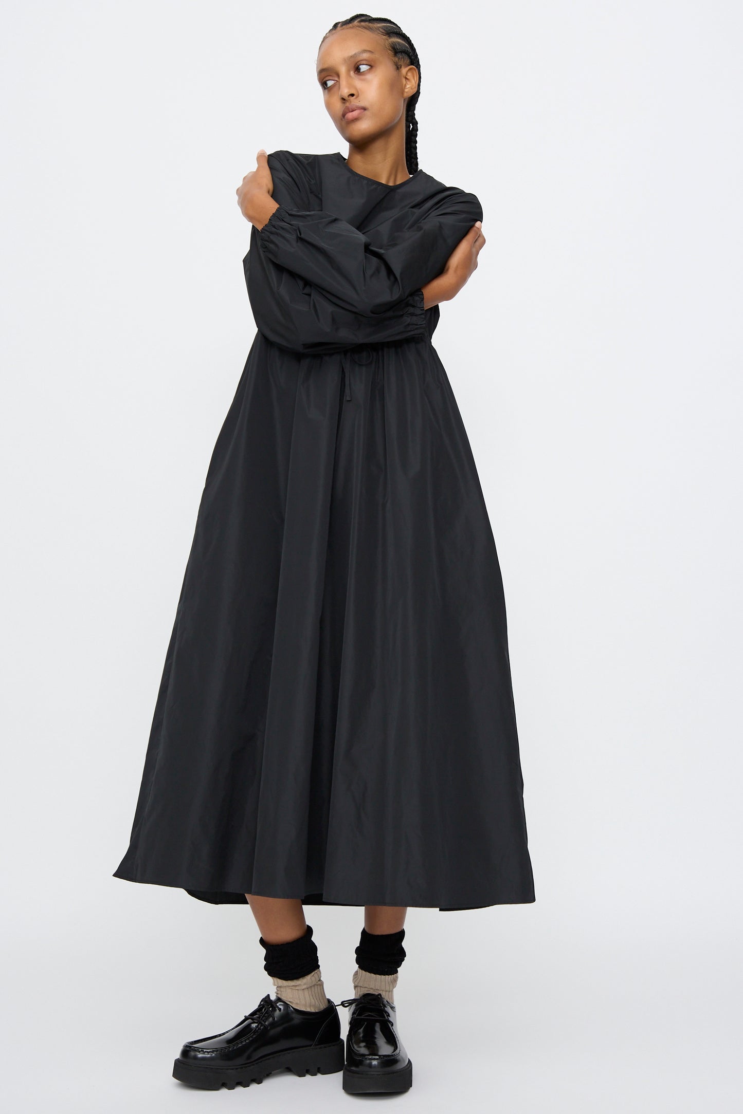 A person with braided hair wears the AVN New Gem Dress in Black, featuring bishop sleeves, paired with black platform shoes and beige socks, standing against a plain white background.