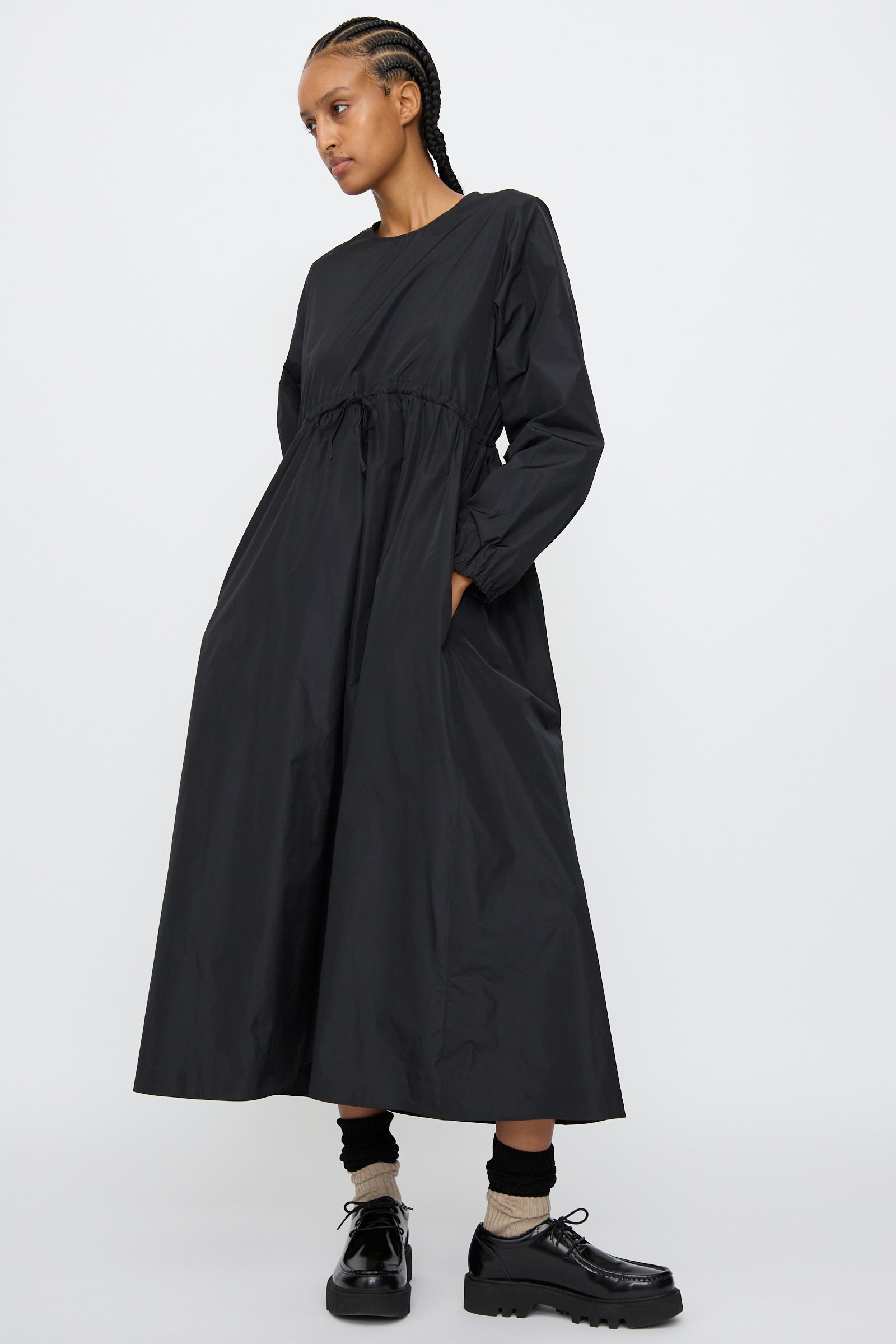 A person with braided hair wears the AVN New Gem Dress in Black, featuring bishop sleeves and side pockets, styled with black shoes and beige socks, standing against a plain light background.