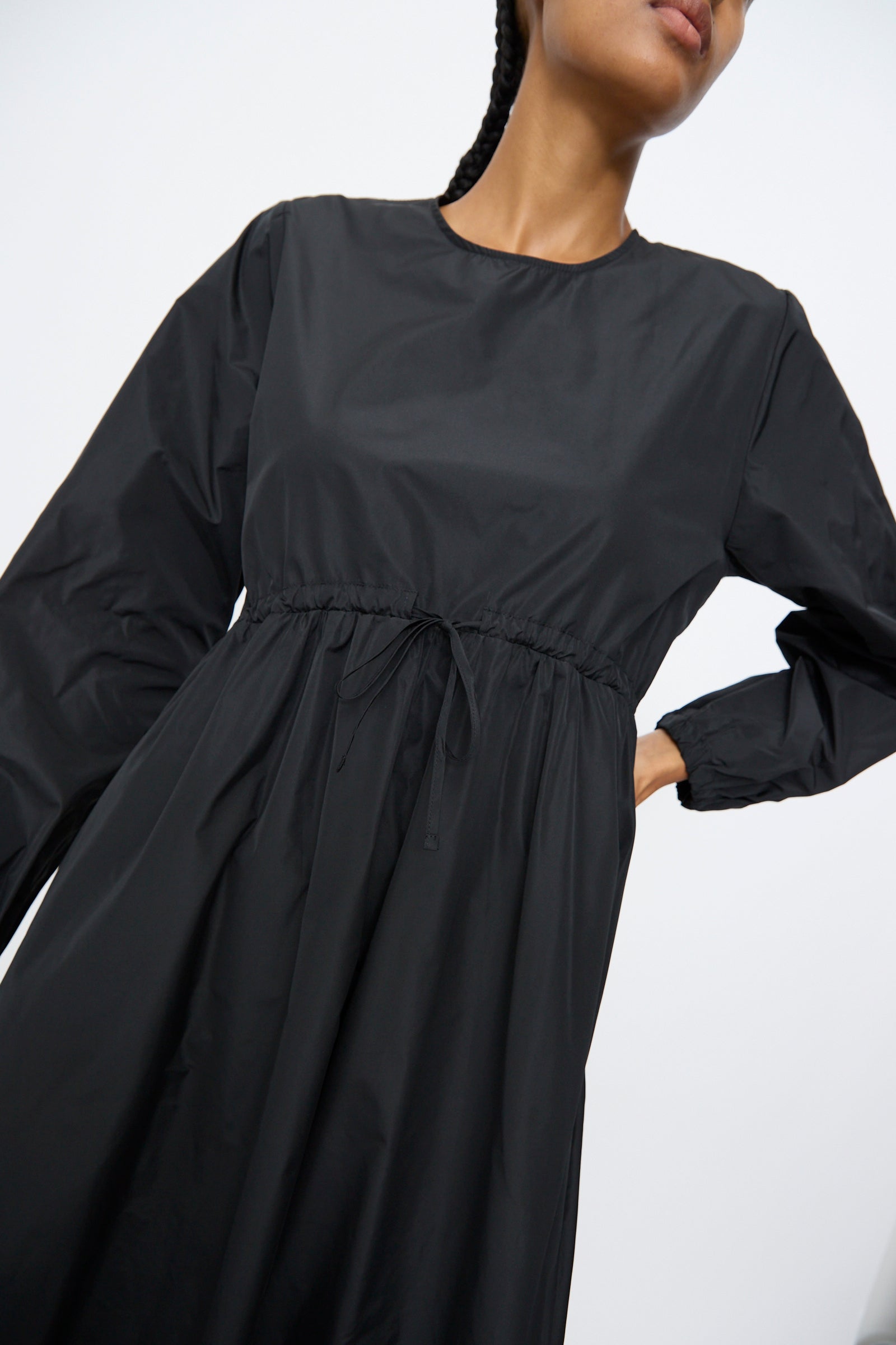 A person models the AVN New Gem Dress in Black, featuring bishop sleeves and an adjustable tie waist, against a plain light background.