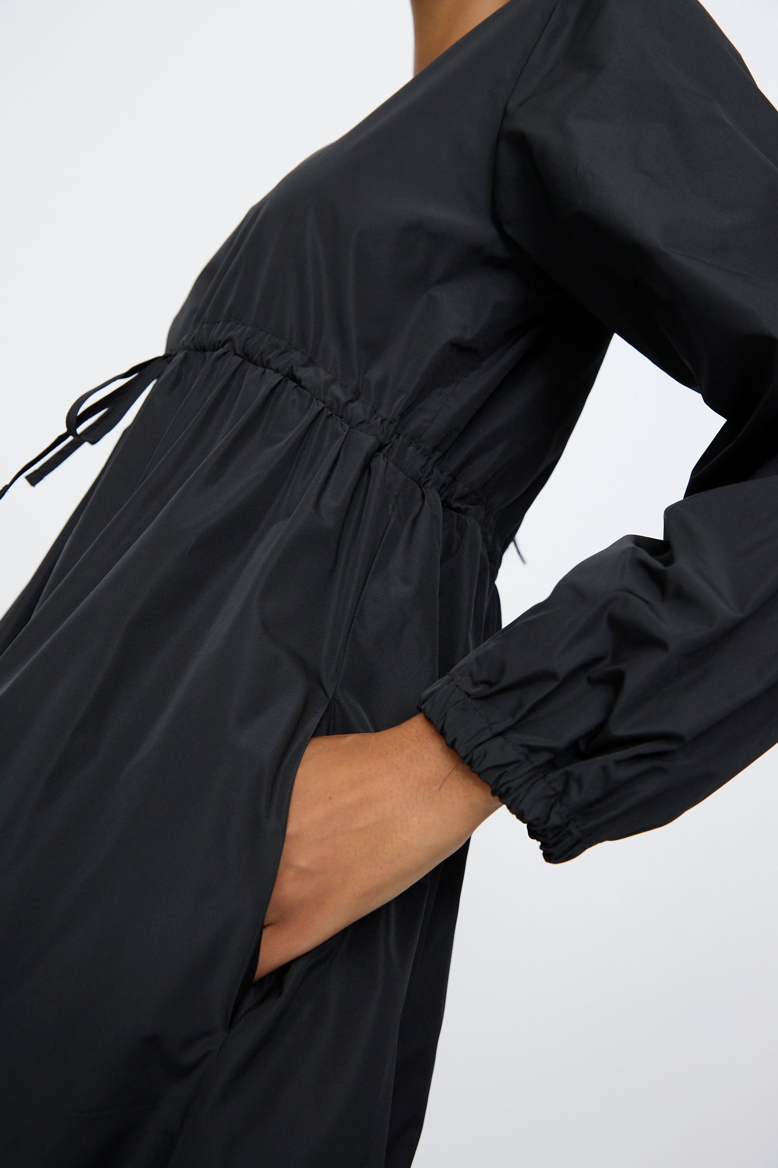 A person wears the AVN New Gem Dress in Black, featuring bishop sleeves, a cinched adjustable tie waist, and one hand in the pocket.