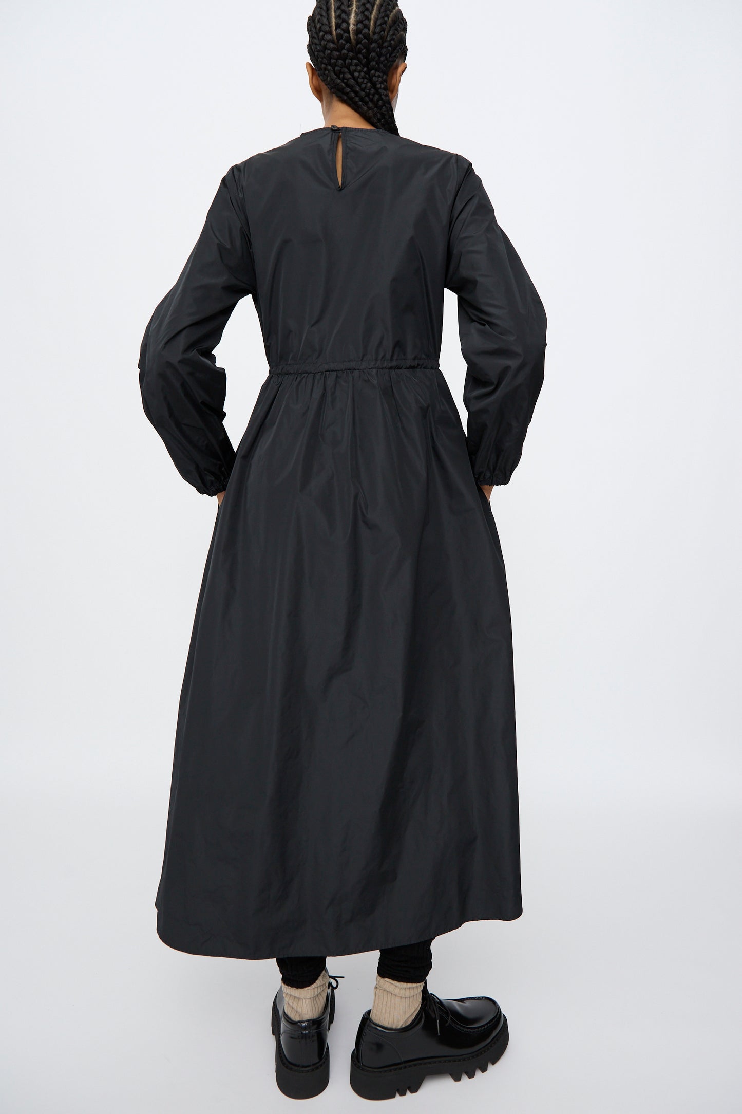 A person stands facing away, wearing the AVN New Gem Dress in Black with bishop sleeves, paired with black shoes and light-colored socks.