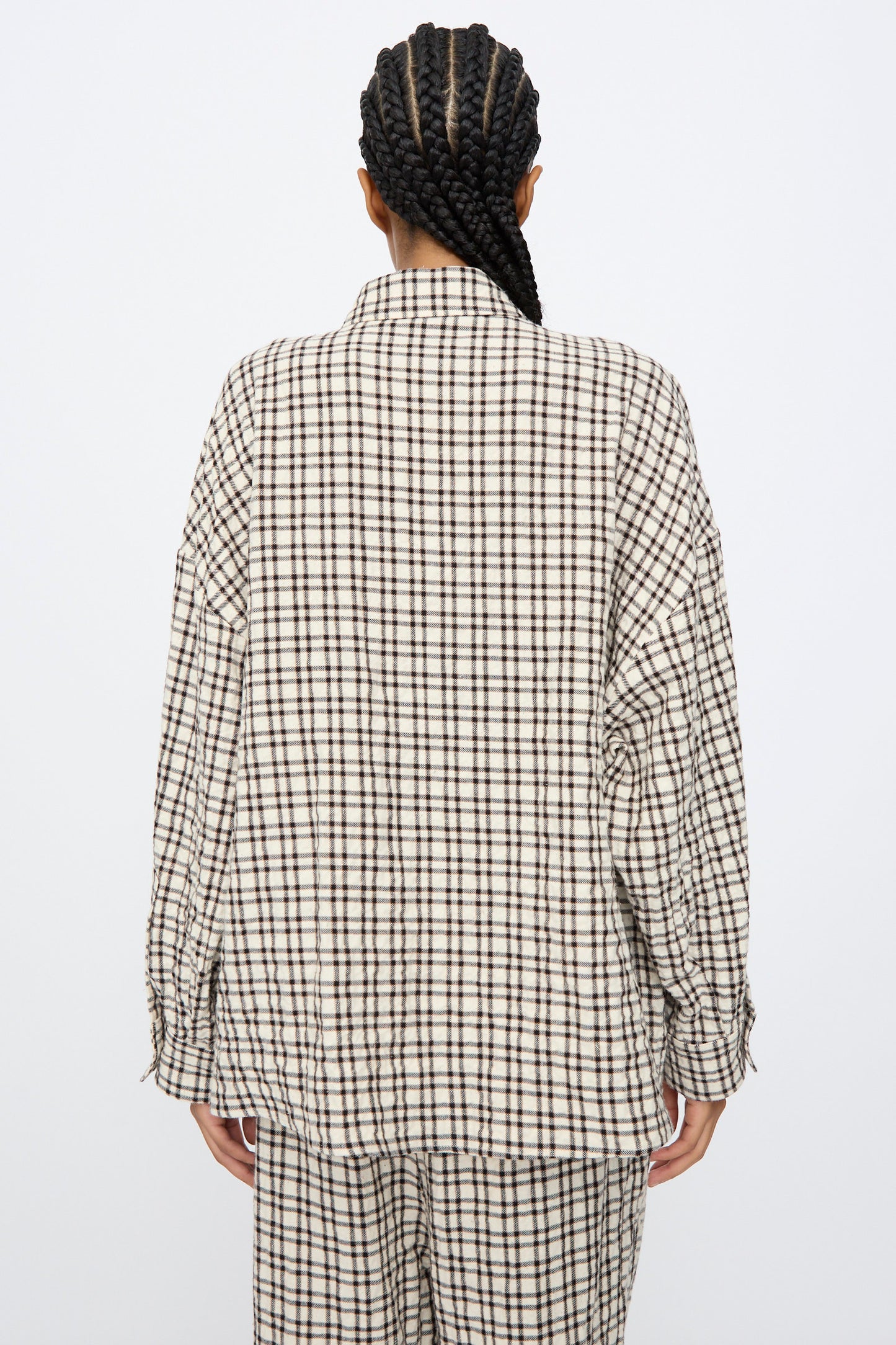 Person with braided hair facing away, wearing AVN's Over Shirt in White and Black Check— a loose-fitting, long sleeve cotton shirt made in Italy—against a plain background.